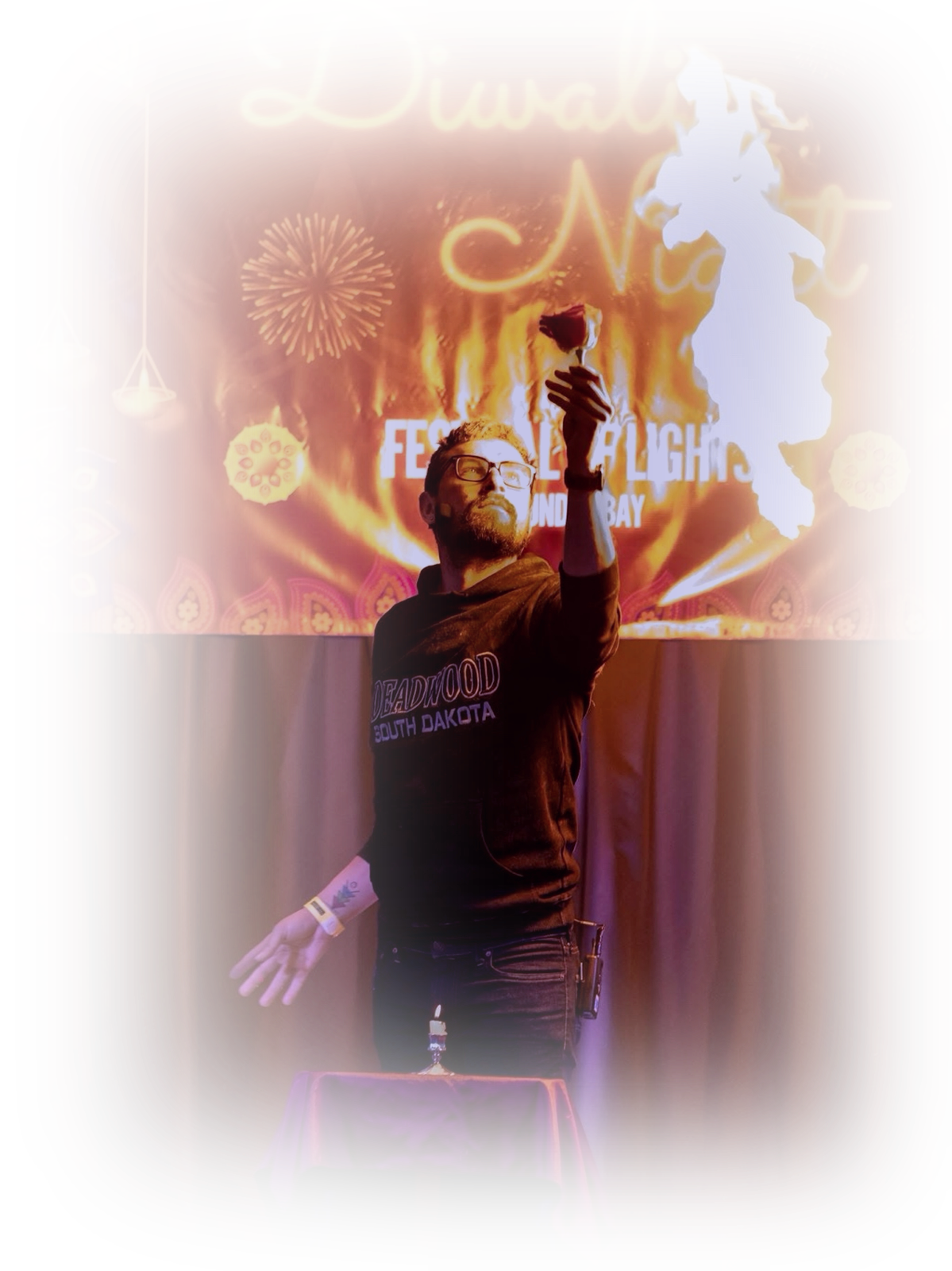 Man with glasses and beard performing a magic trick at a Diwali celebration, holding a rose and a candle, with a backdrop featuring Diwali-themed decorations, fireworks, and the text 'Diwali Nat'.