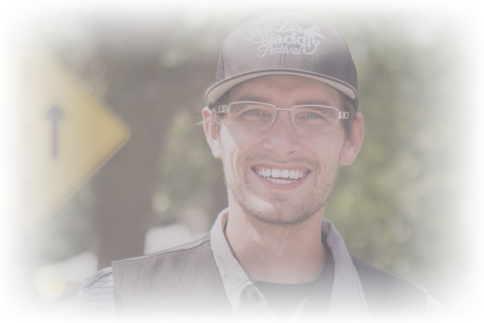 A smiling man wearing glasses and a cap with the logo 'Double Lake Blackfly Festival', standing outdoors with trees in the background.