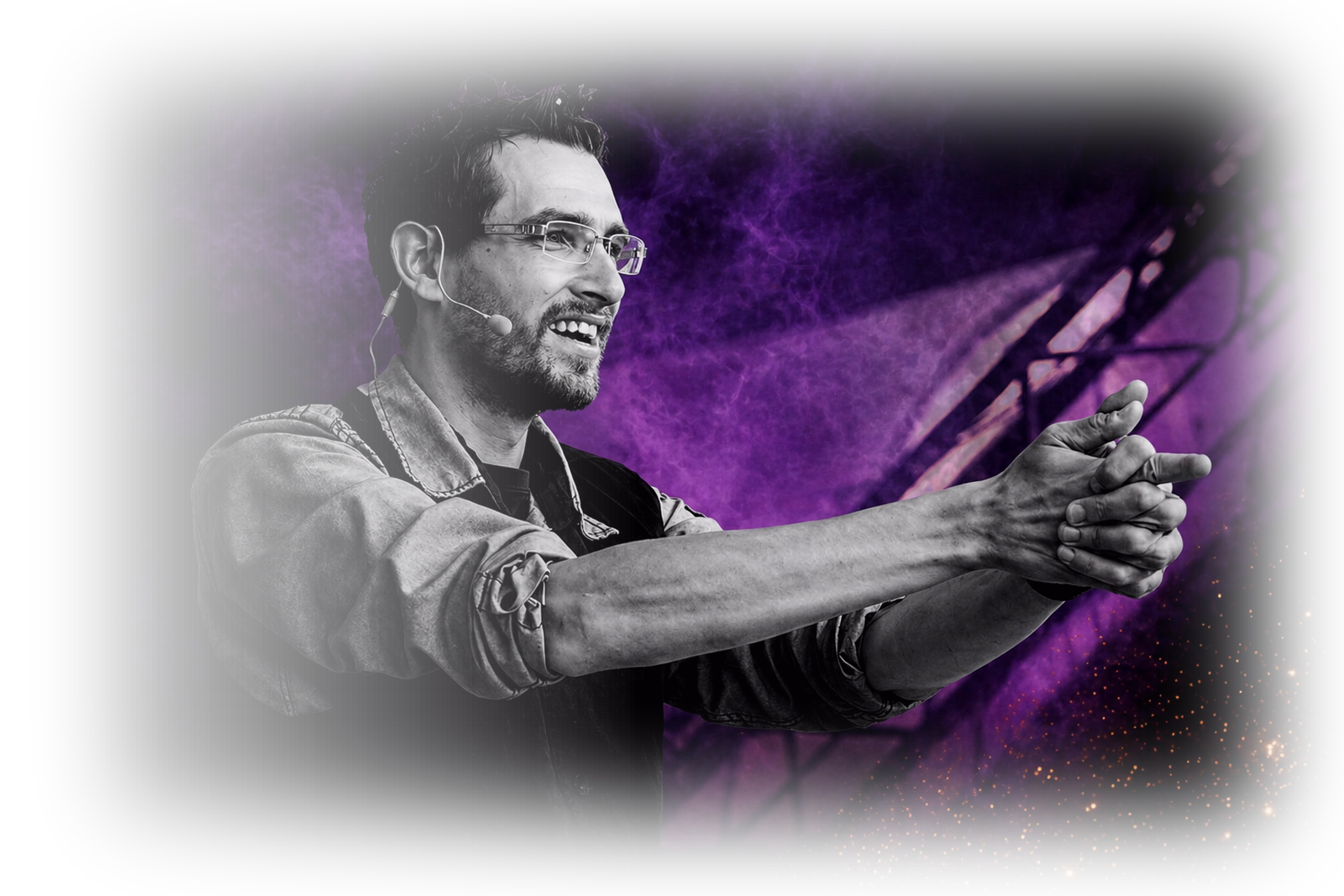 A man wearing glasses and a microphone headset, with tattoos on his arm, is smiling and gesturing with his hands against a background of purple smoke, sparks, and stage lights.