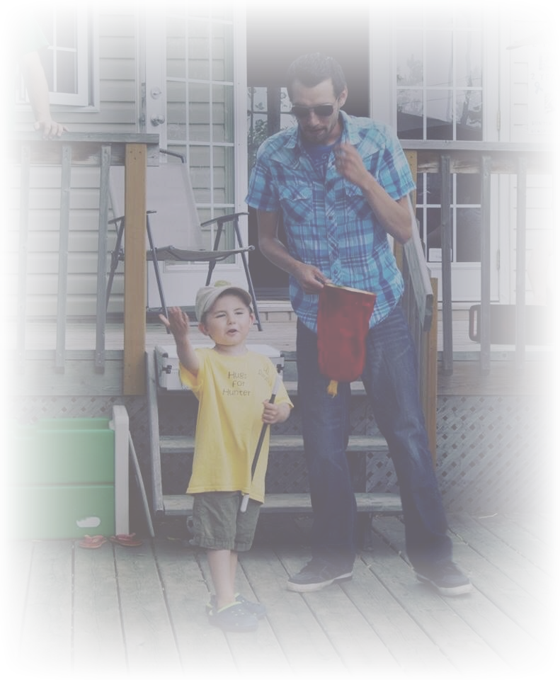A young boy wearing a yellow shirt and shorts holding a stick, raising his hand, standing on a wooden deck with an adult man in sunglasses and a blue plaid shirt beside him. In the background, there are chairs, a house with siding, and a sliding glass door.
