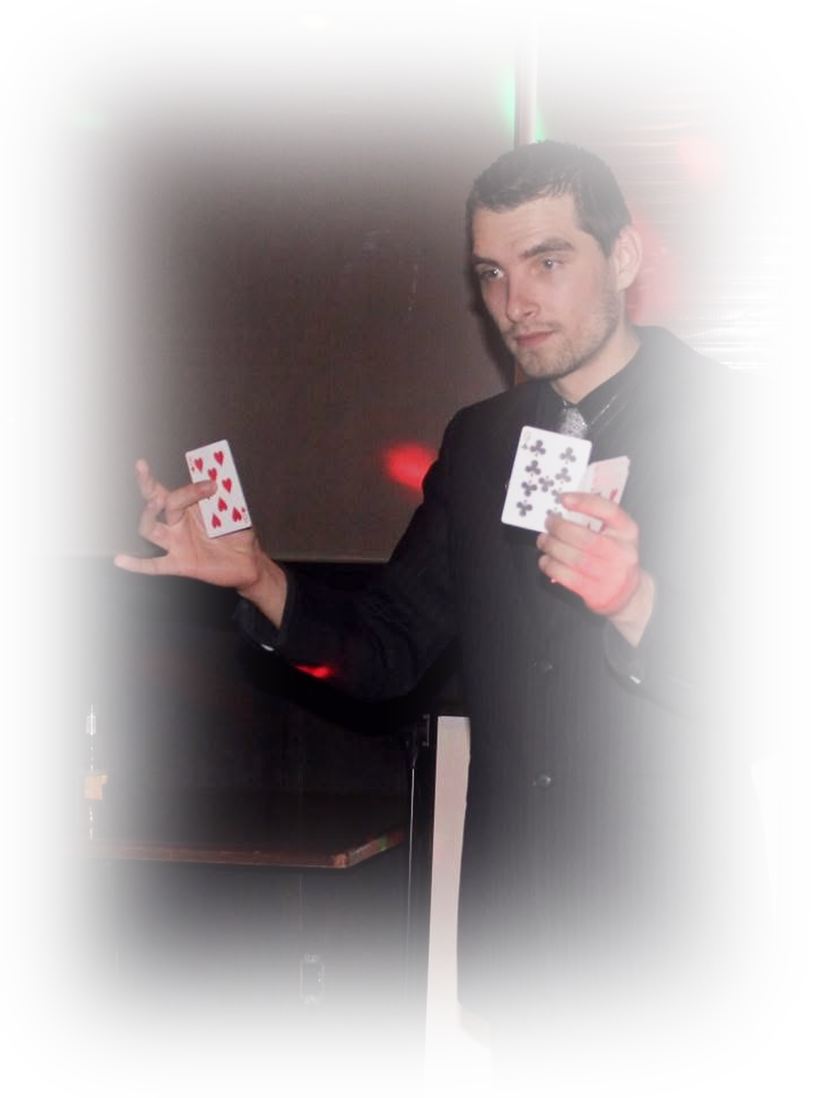 A magician in a black suit performing a card trick with two playing cards in a dimly lit room with colorful lights reflected on the wall.