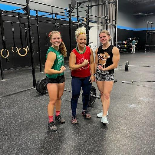 Three women in athletic wear at a gym, standing in front of gym equipment, smiling and posing for the camera.