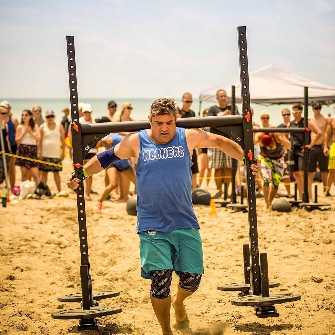 A man participating in a strongman event on a sandy beach, lifting a heavy frame, with an audience watching under a tent in the background.