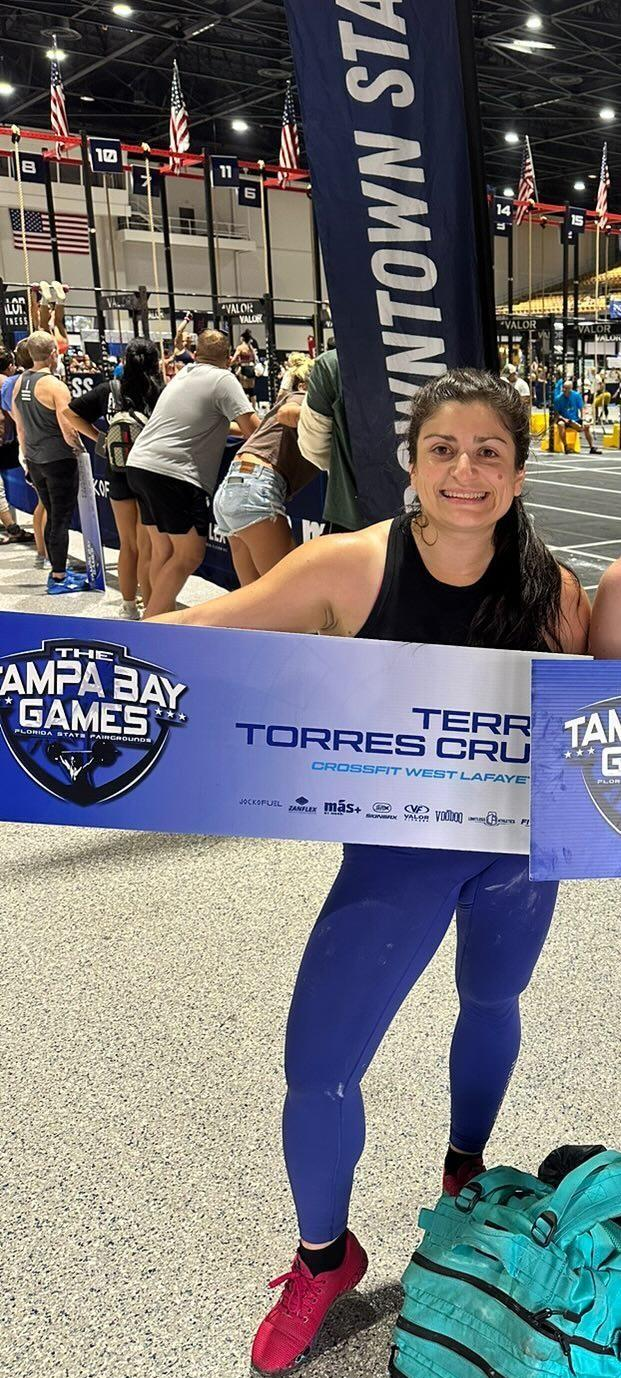 A woman holding a banner at a fitness competition event called The Tampa Bay Games, with an indoor gym setting in the background.