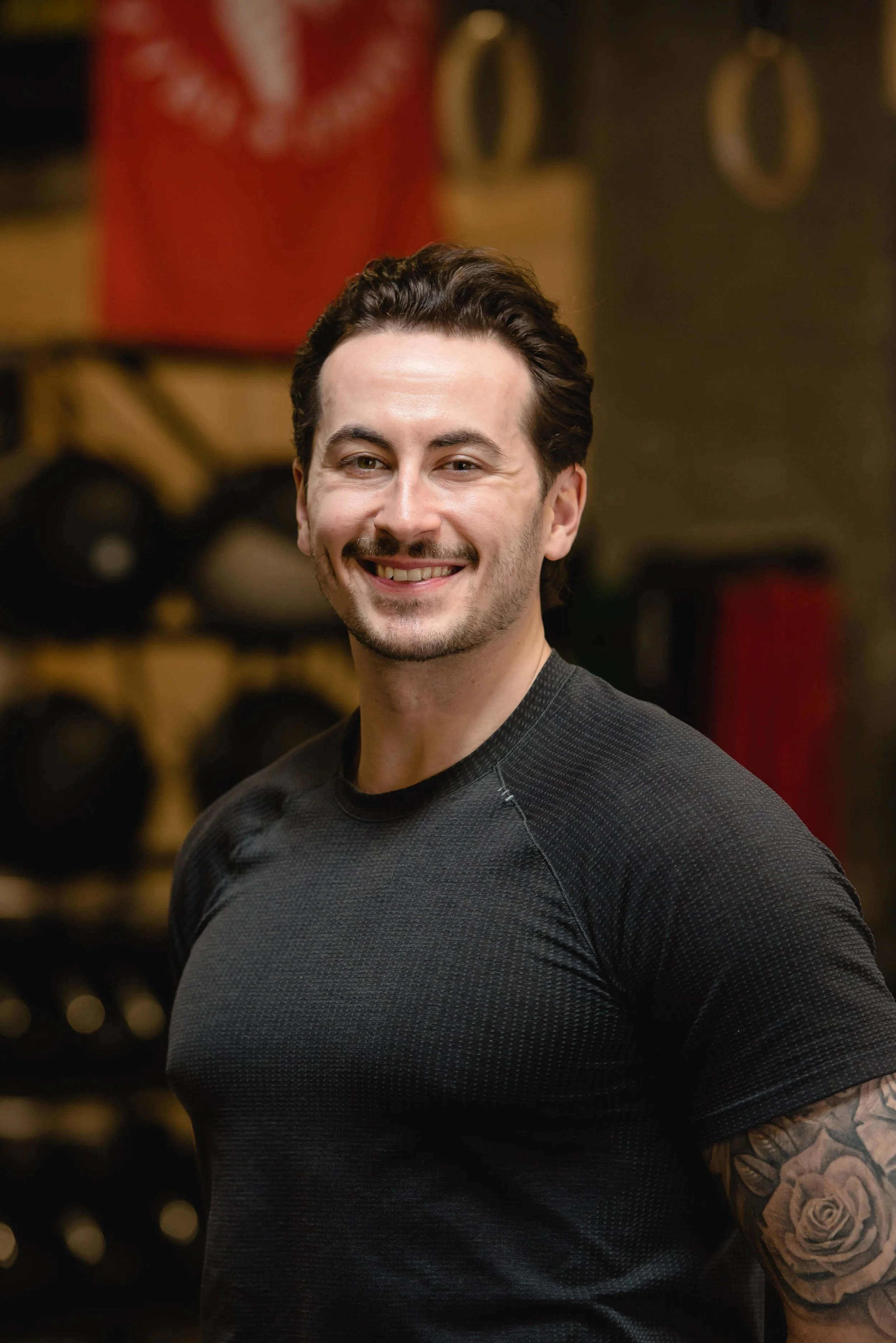 A young man with dark hair, a light beard, and tattoos on his arm, smiling and wearing a black athletic shirt, standing in a gym with blurred weights in the background.