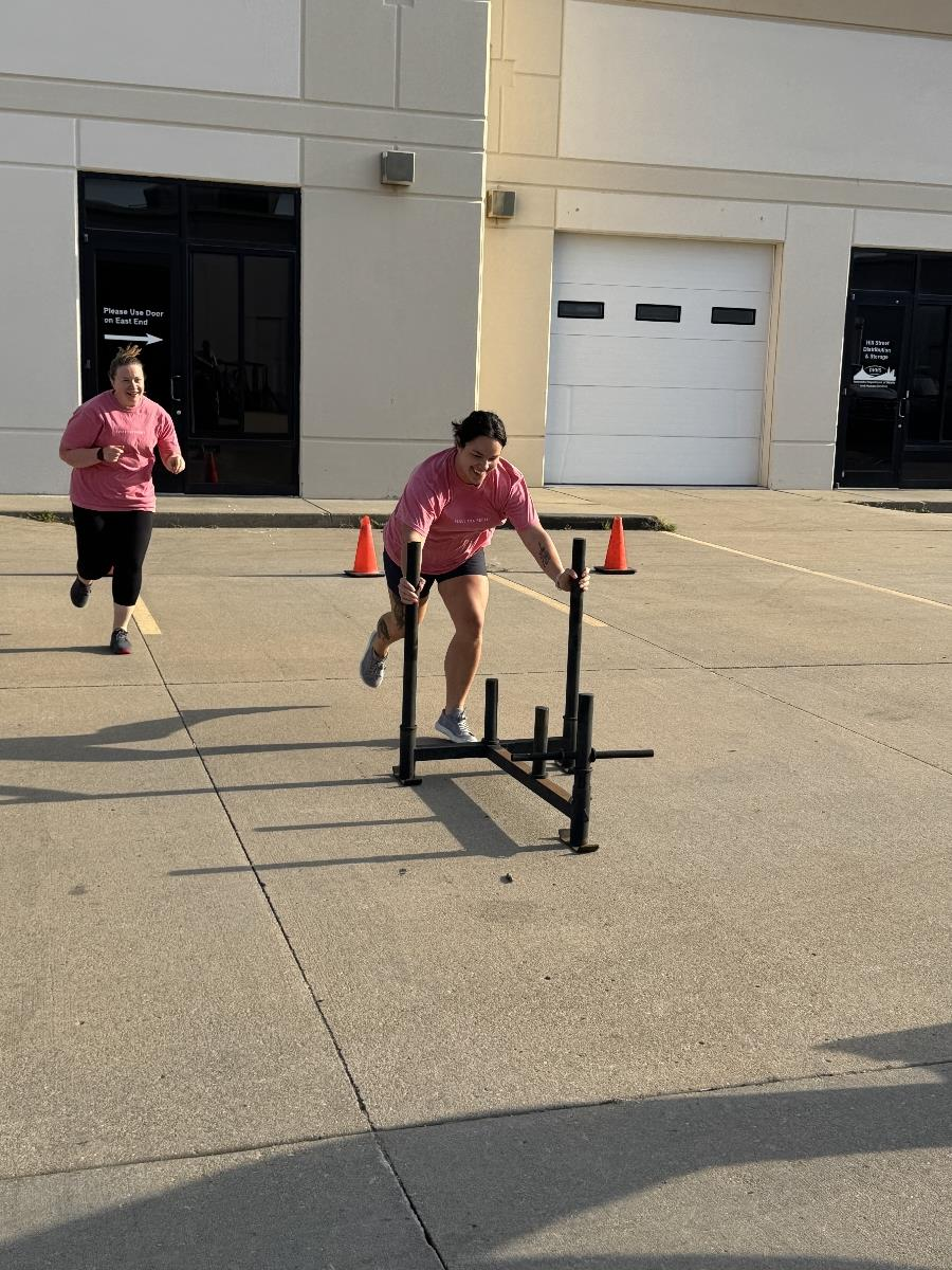 Two women in pink shirts participating in an outdoor fitness activity. One woman is pushing a sled, and another woman is running behind her. Orange cones mark the area near a building.