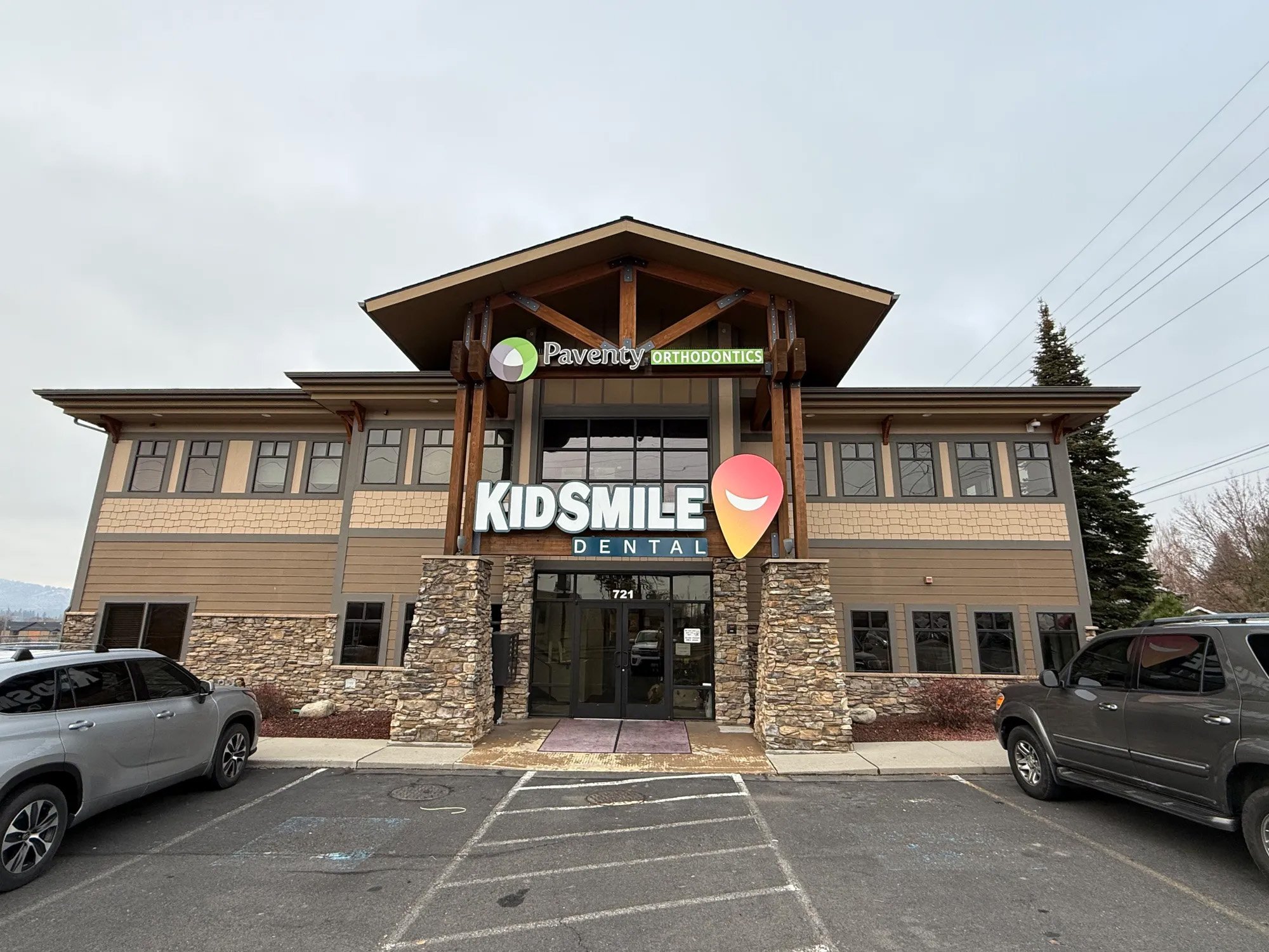 A two-story building housing Kidsmile Dental, with a sign for Pavenyt Orthodontics above the entrance. The building features stone accents on the lower part, brown siding, and large windows. There are parked cars in front and a parking lot with strip
