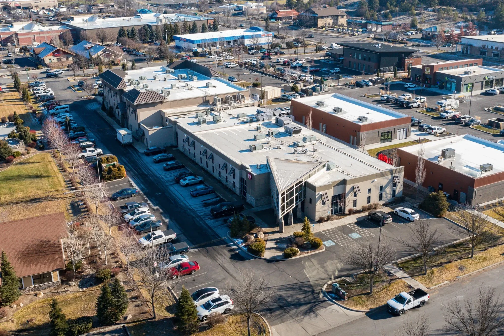 An aerial view of a shopping center with multiple stores, parking lots with many parked cars, and surrounding trees and buildings.