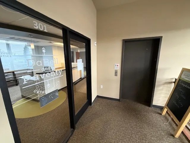 Hallway with elevator doors, windowed office entrance labeled 'D. Bell and Pavenity Orthodontics,' and a small chalkboard sign on an easel.