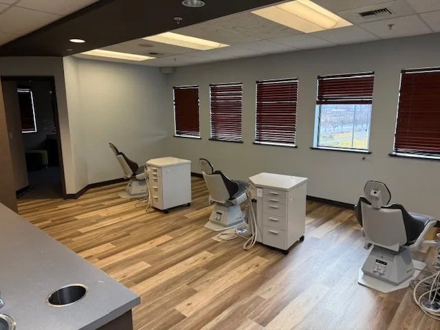 Empty dental clinic treatment room with four dental chairs, small white cabinets, and large windows with closed blinds.