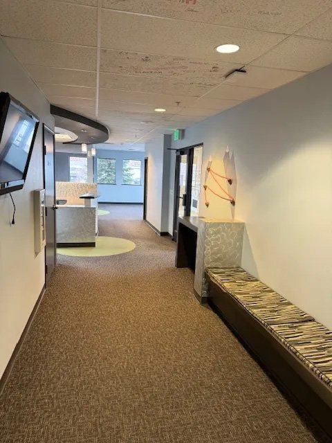 Empty hospital or clinic corridor with a bench, wall-mounted TV, and a reception or information desk in the background.