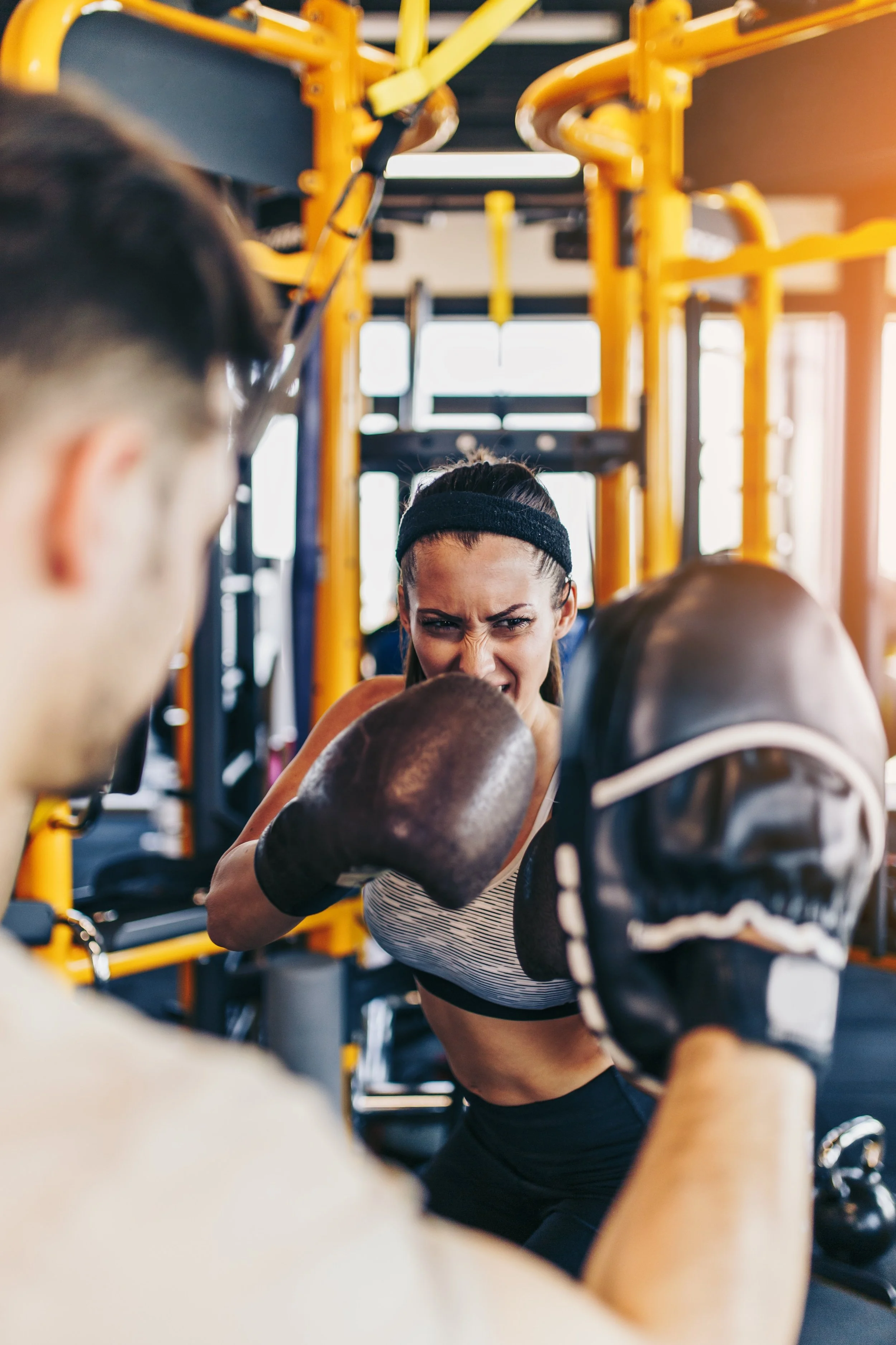A woman wearing boxing gloves training with a coach in a gym with yellow exercise equipment.