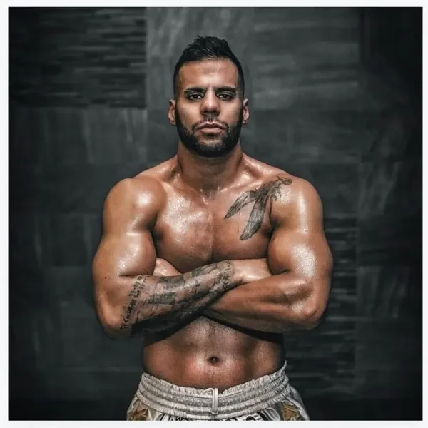 A muscular man with tattoos crossing his arms, standing shirtless against a dark background.