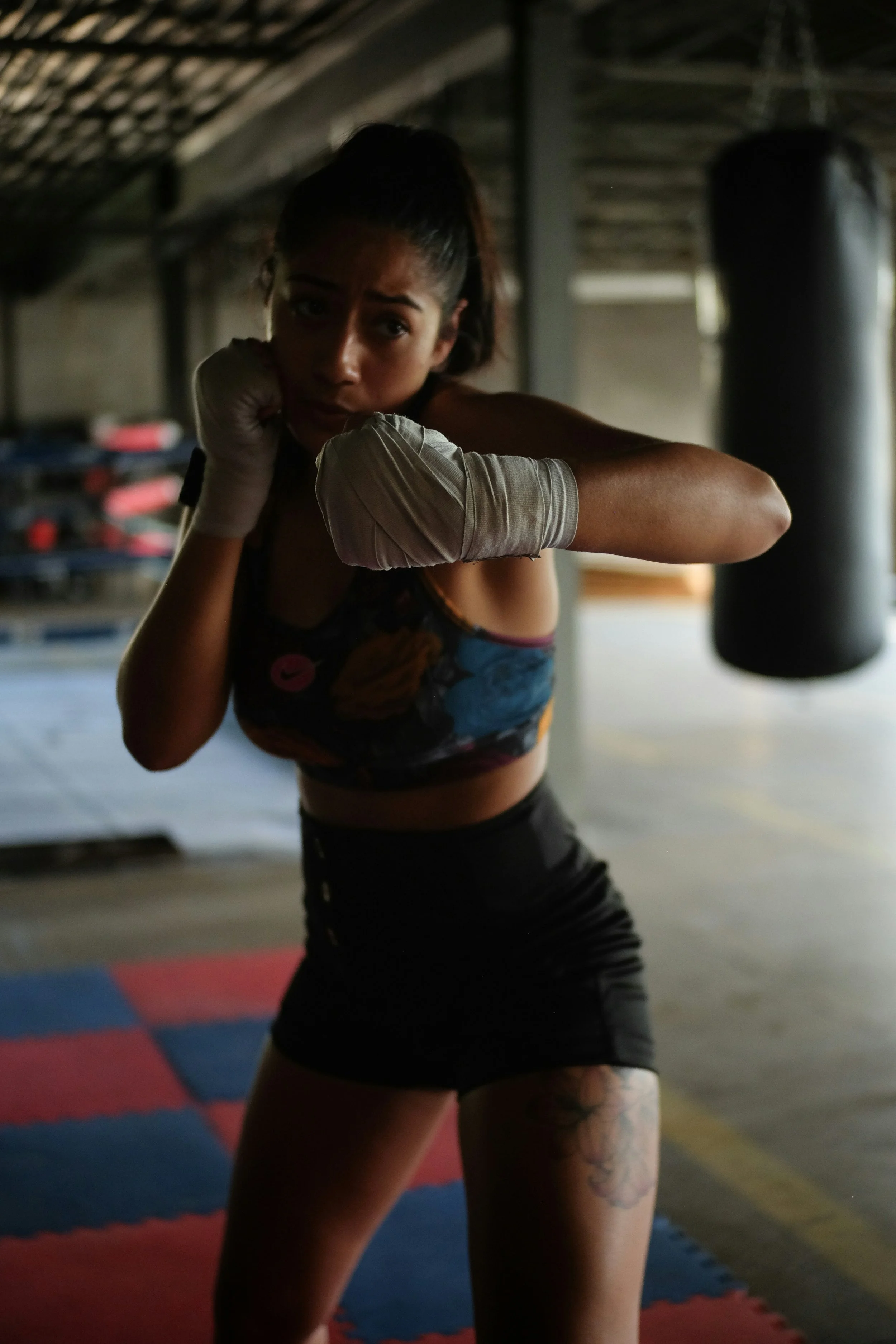 A woman practicing boxing in a gym, wearing gloves, a sports bra, and shorts, with a punching bag in the background.