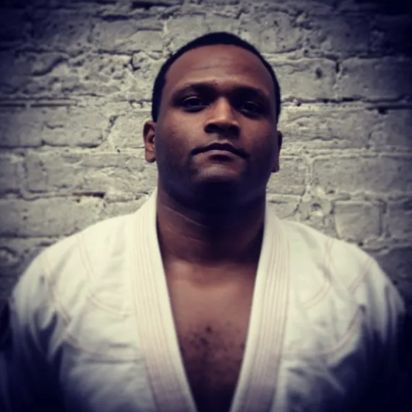 A man with short dark hair and medium skin tone wearing a white martial arts gi in front of a light-colored brick wall.
