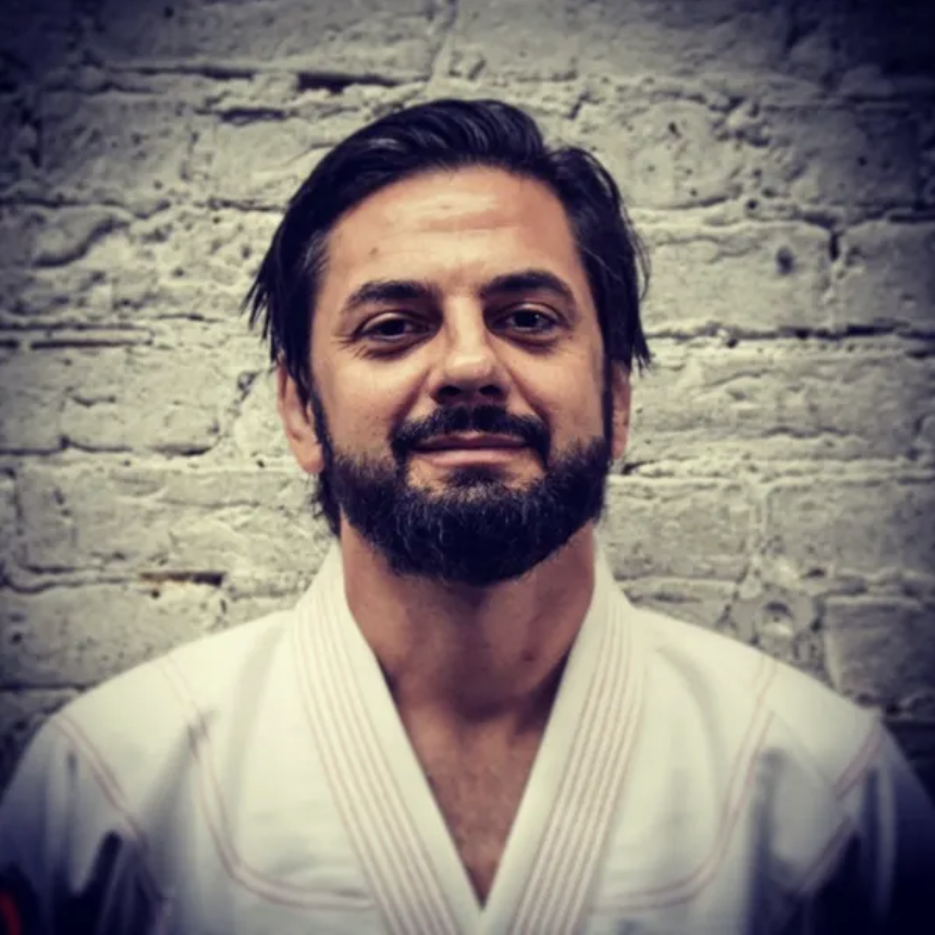 A man with dark hair, a beard, and a mustache, wearing a white martial arts uniform, standing in front of a brick wall.