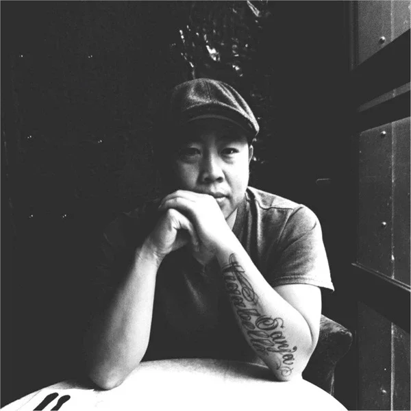 A person sitting at a table with their chin resting on their hands, wearing a flat cap and a short-sleeved shirt, with a tattoo on their forearm, in a dimly lit environment.