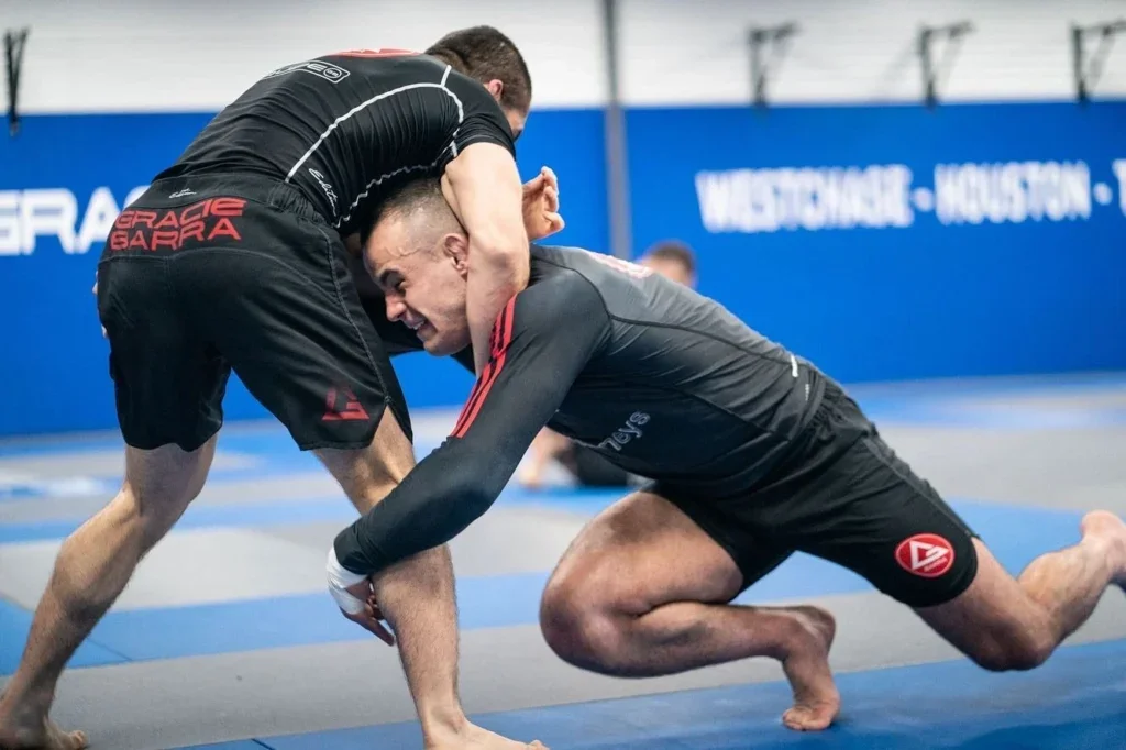 Two men are practicing Brazilian Jiu-Jitsu on mats, one in a black rash guard and the other in a gray rash guard, engaged in a grappling position.