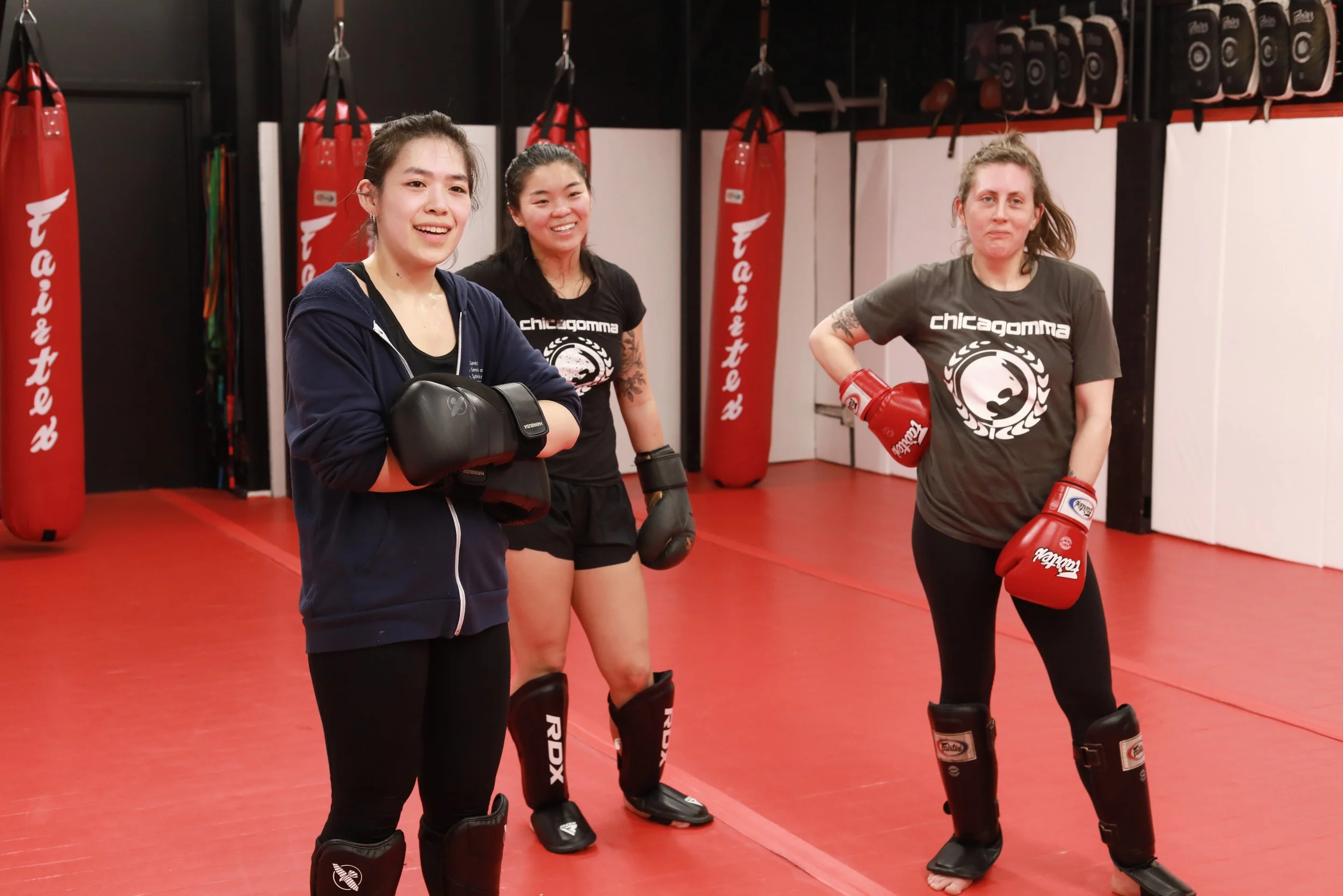 Three women wearing fight gear standing in a martial arts gym with punching bags and training equipment visible in the background.