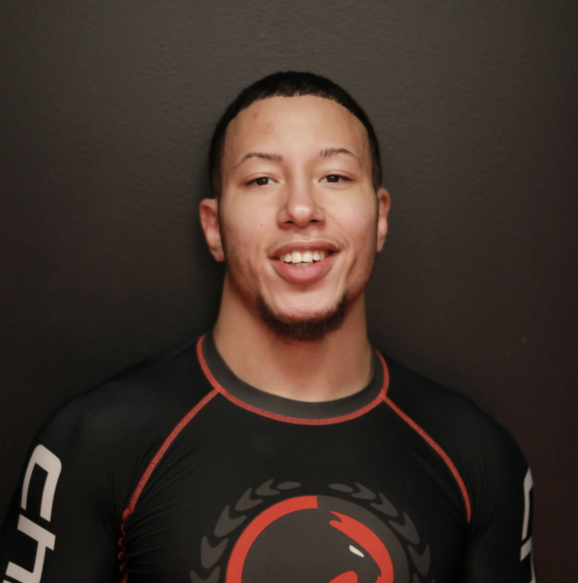 Young man with short dark hair and a goatee smiling, wearing a black athletic shirt with red and white accents, standing against a dark background.
