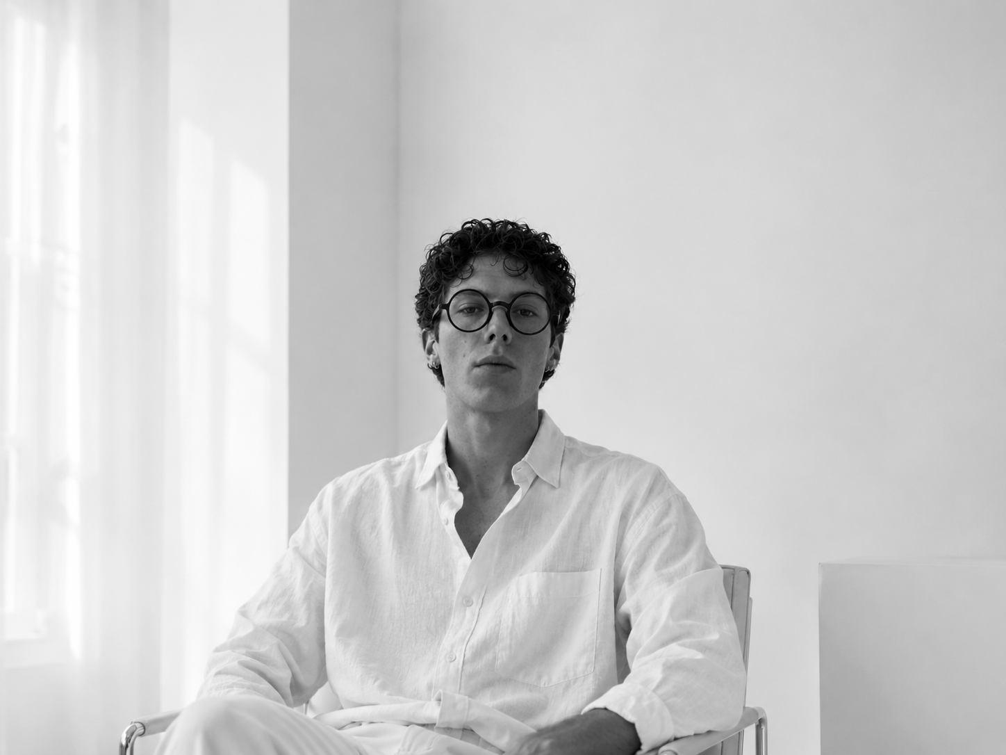 Black and white photo of a young person with curly hair and glasses, leaning on a chair with one hand on the backrest, in front of a plain wall.
