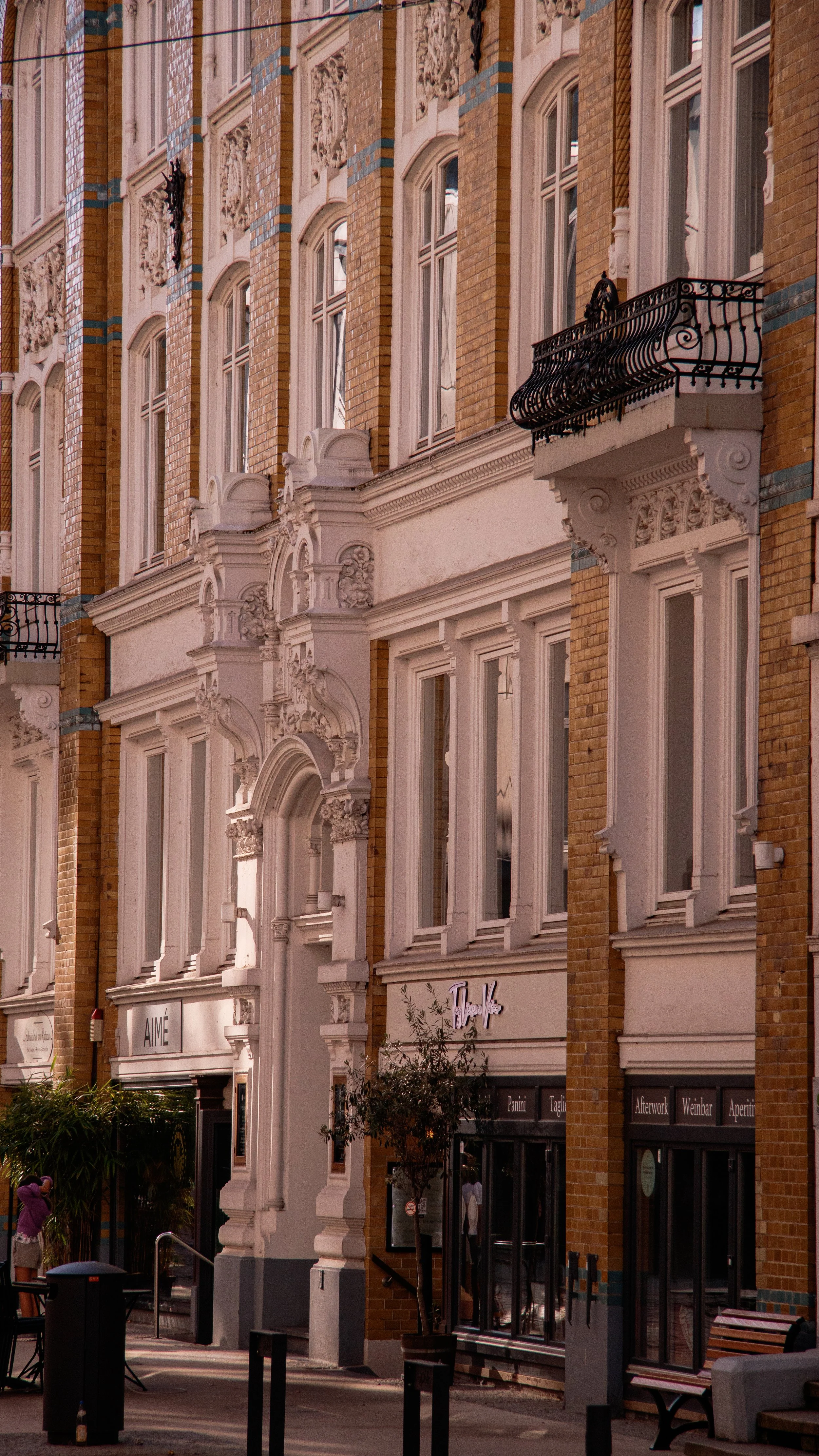 Fassade eines mehrstöckigen, historischen Gebäudes mit großen Fenstern und dekorativen weißen Elementen, kleine Balkone mit schwarzen Geländern, Ladenflächen im Erdgeschoss, Menschen und Bänke auf dem Gehweg.