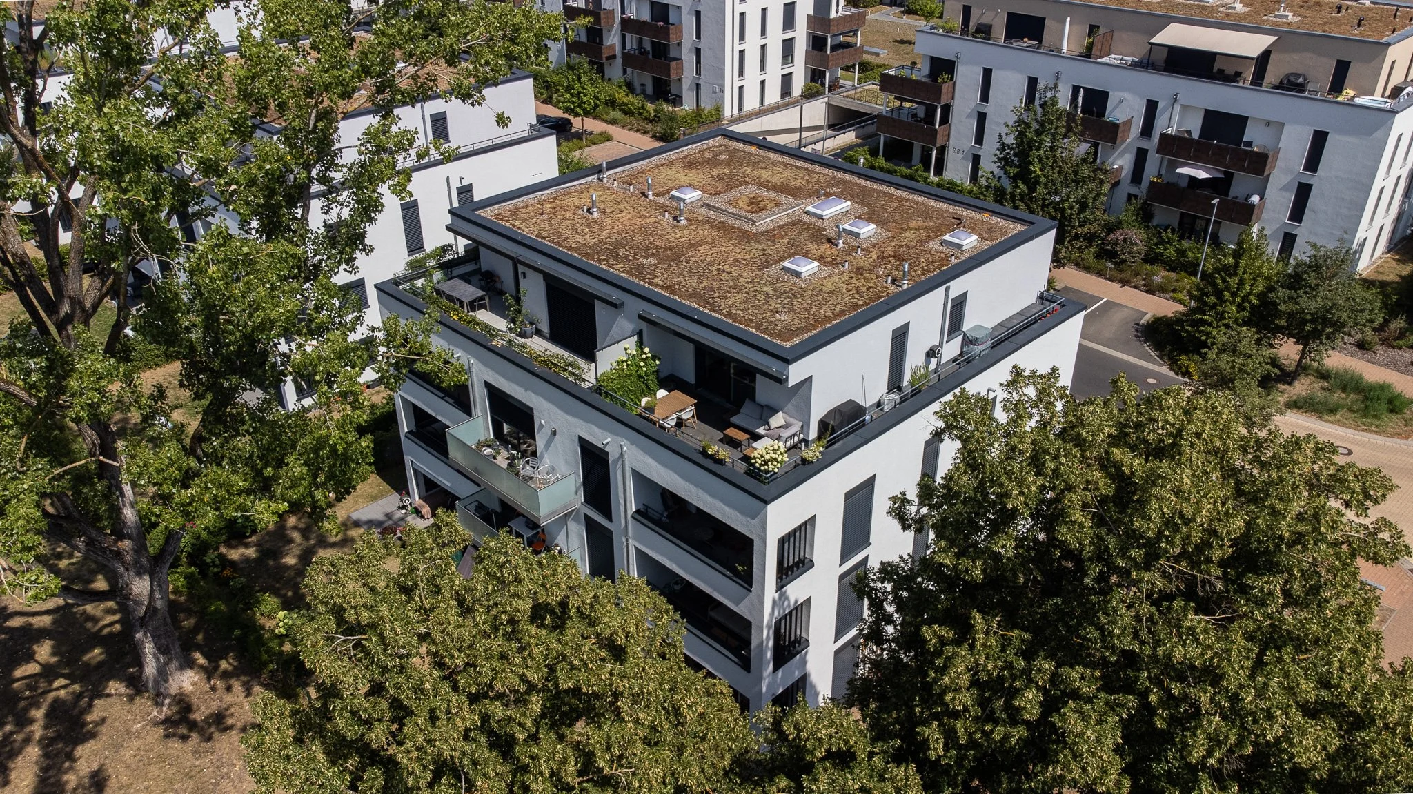 Luftaufnahme eines modernen Mehrfamilienhauses mit Dachterrasse und umgebenen Grünflächen in einer städtischen Gegend.