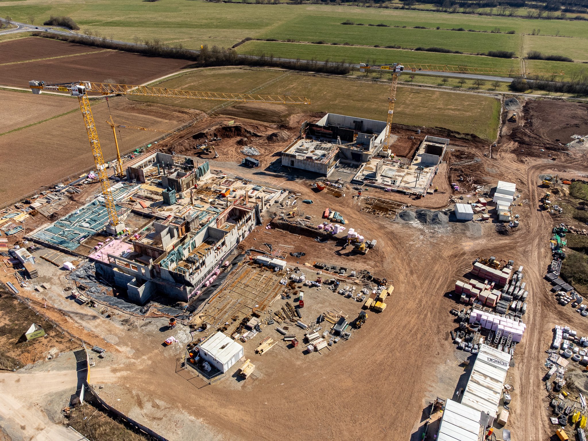 Eine Baustelle mit mehreren Baukränen und Bauarbeiten in einem ländlichen Gebiet mit Feldern im Hintergrund.