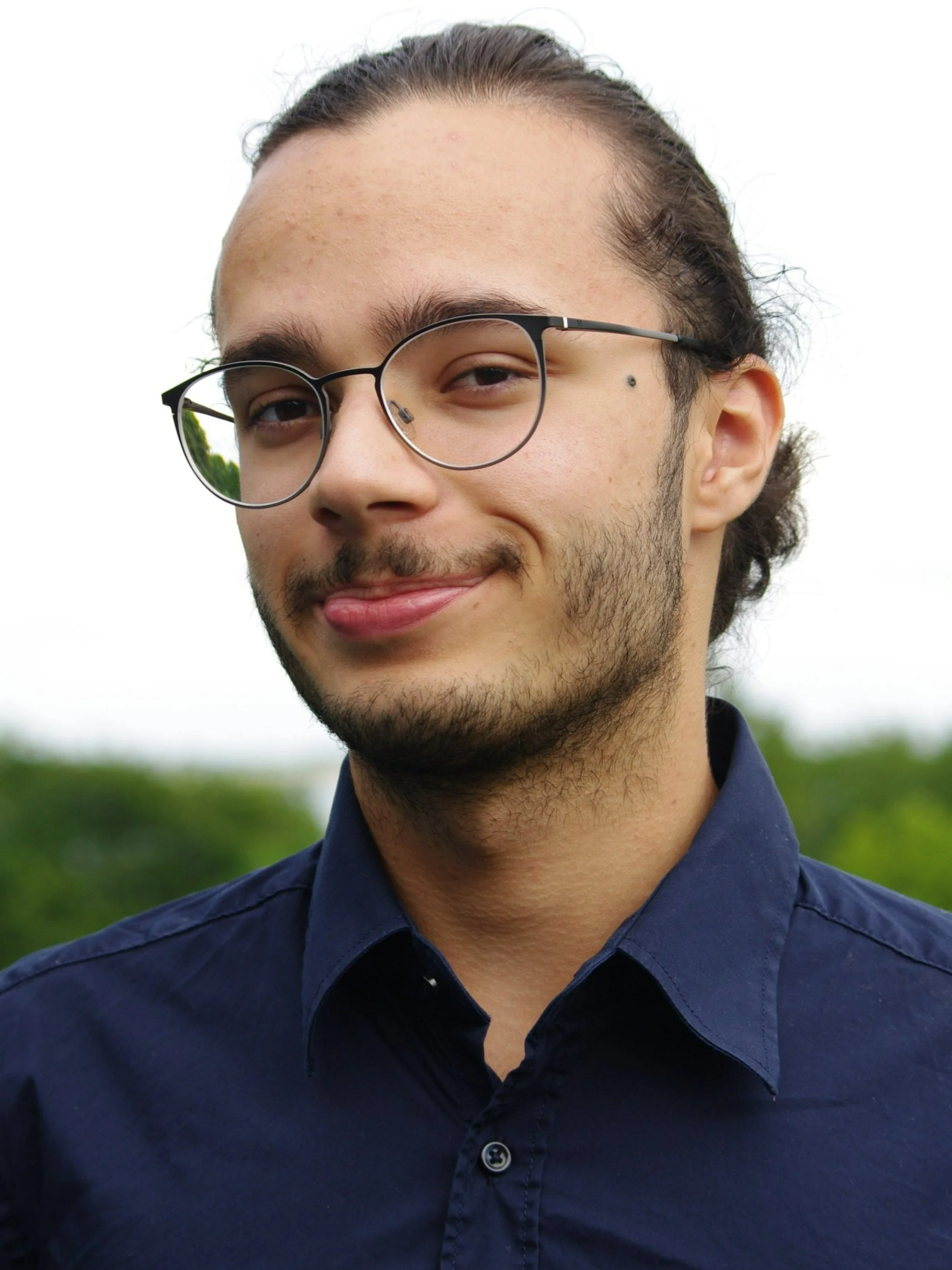 Junger, sympathischer Mann mit Brille, dunklen Haare, leichten Bart, lächelt, trägt dunkles Hemd, im Freien mit grüner Landschaft im Hintergrund.
