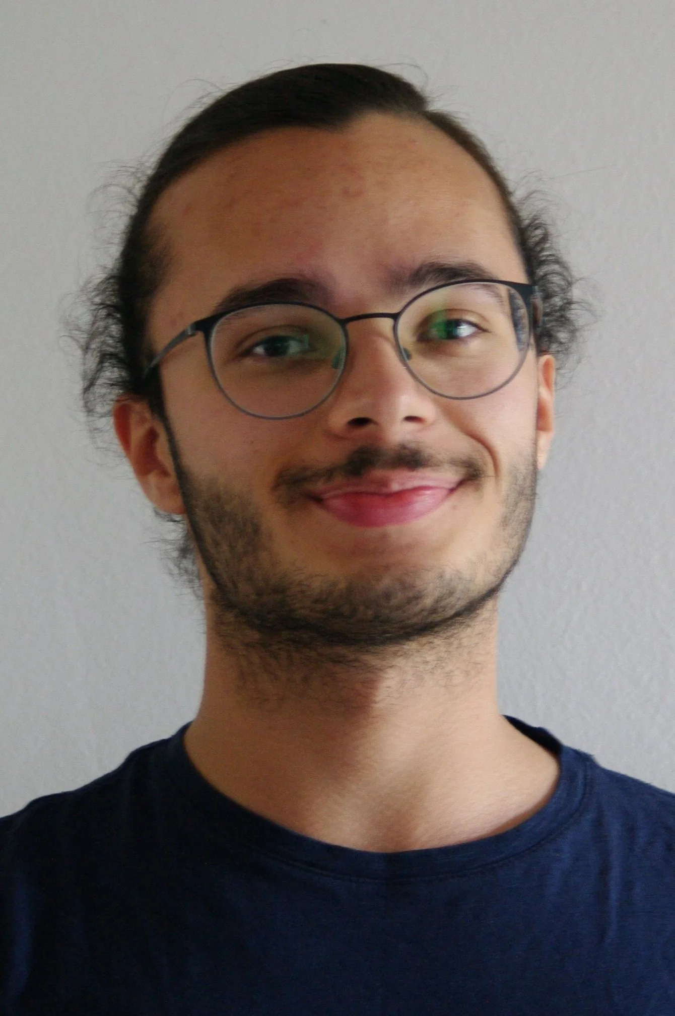 Porträt eines jungen Mannes mit Brille, dunklem Haar, Bart und einem freundlichen Lächeln, vor einem hellen Hintergrund.