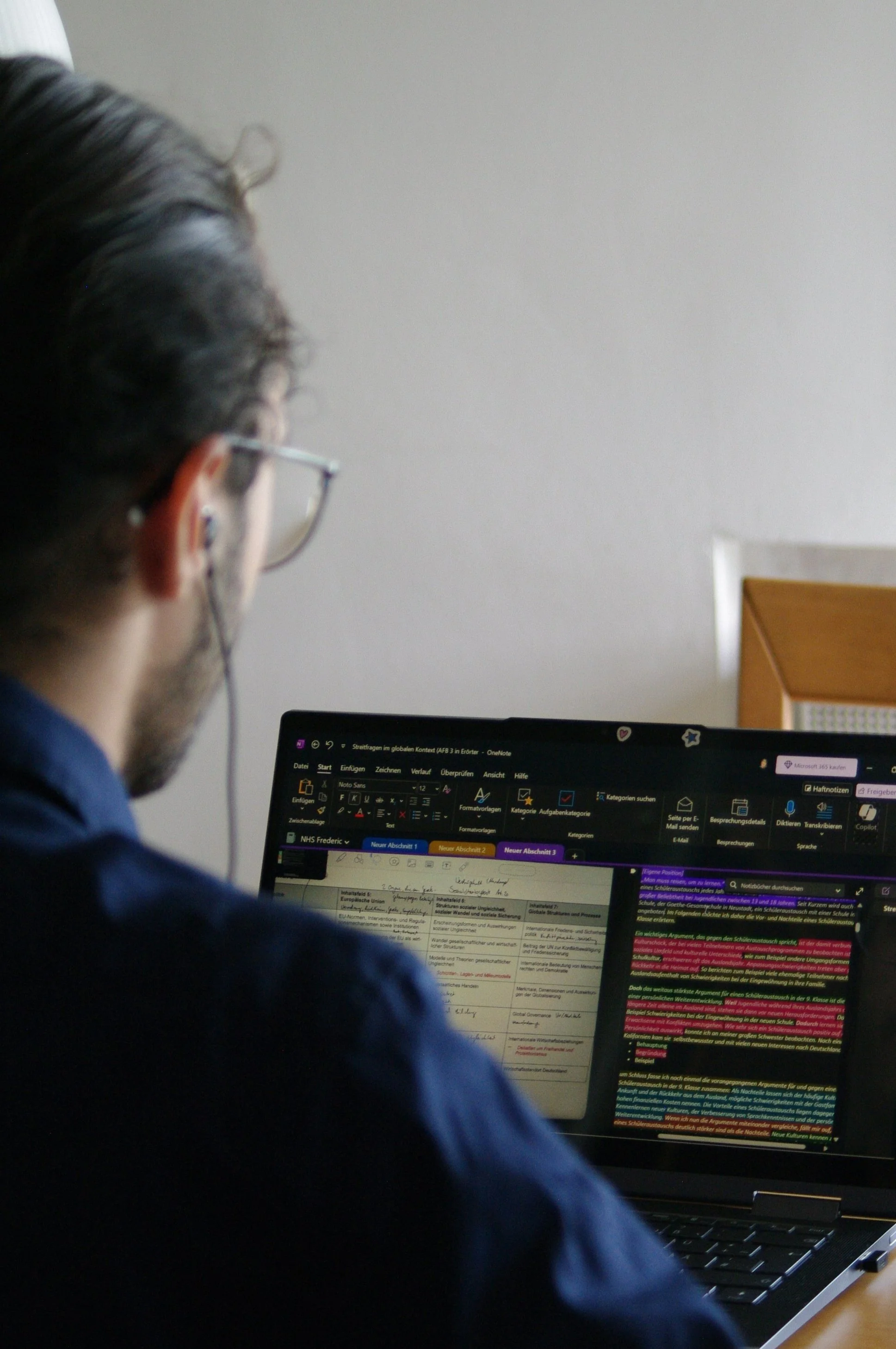 Mann arbeitet an einem Laptop mit offenen Dokumenten und farblich markierten Texten, trägt Brille und hat dunkle Haare.