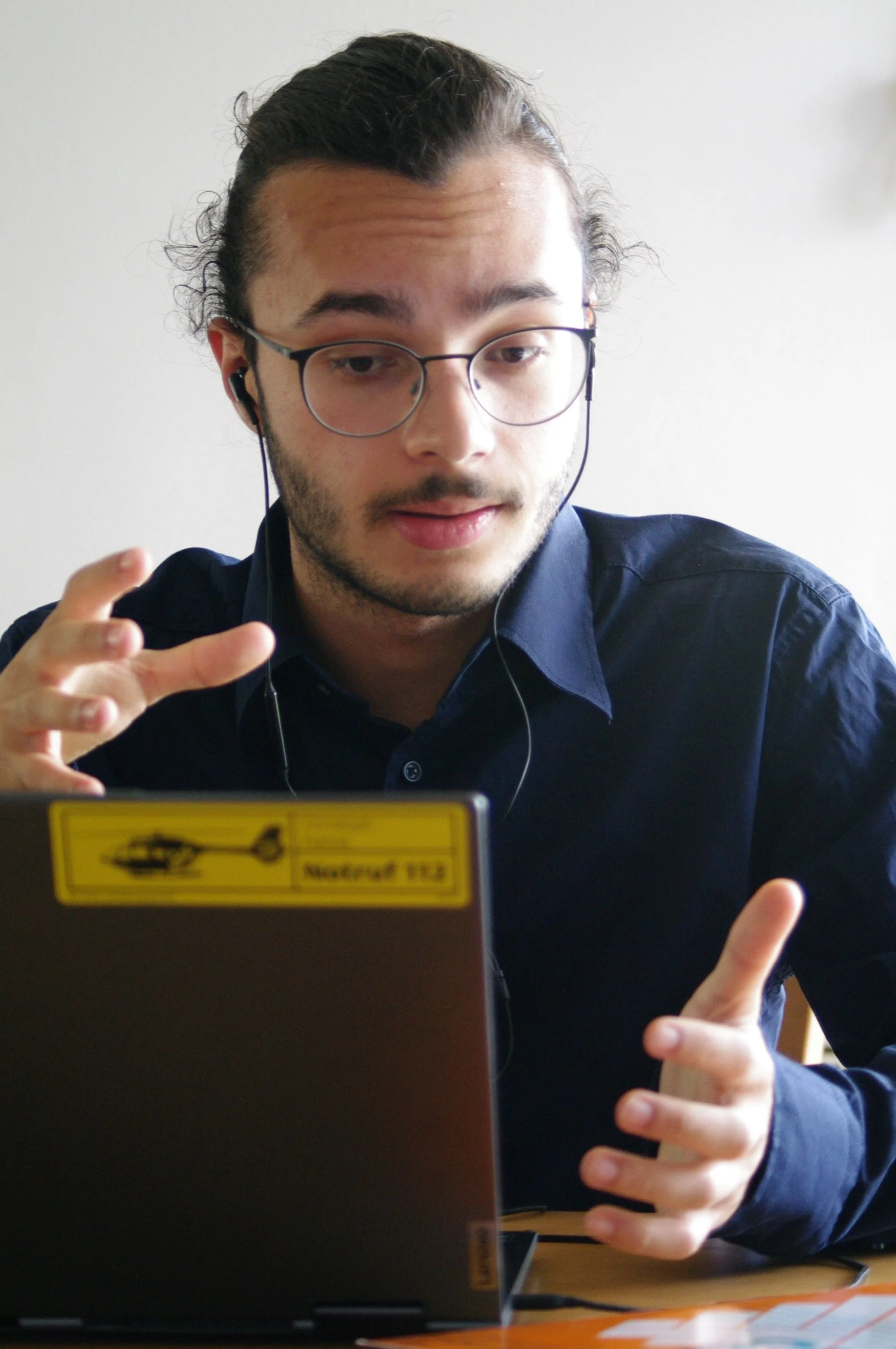 Junger Mann mit Brille und dunklem Hemd spricht während eines Nachhilfe Online Meetings, sitzt an einem Tisch vor einem Laptop.