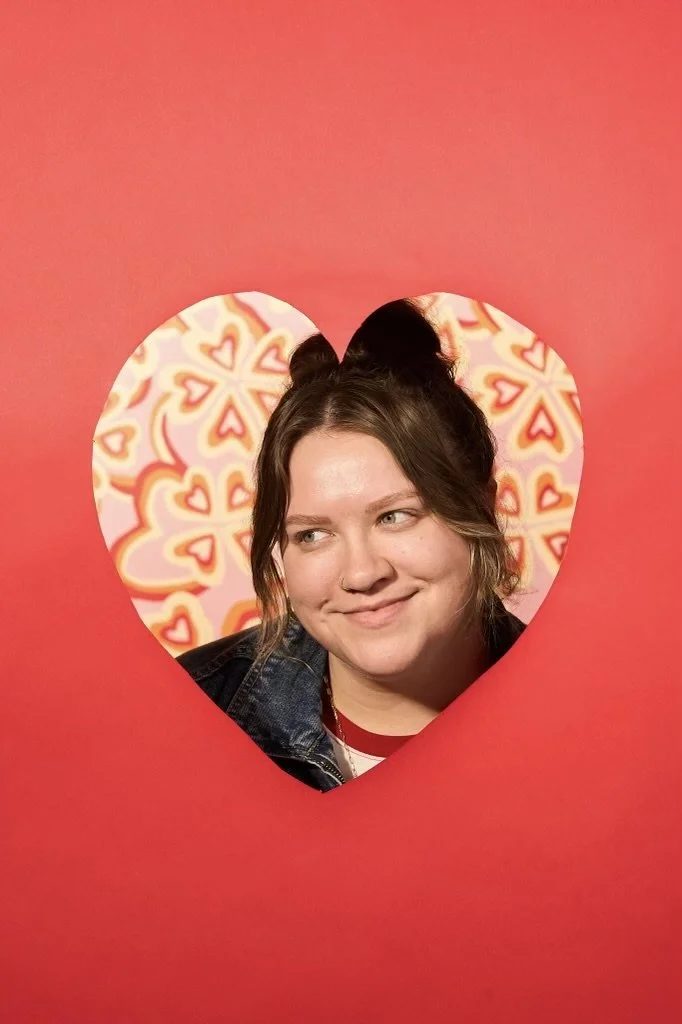 A woman looking through a heart-shaped cutout in a red poster with a background of multicolored heart patterns.