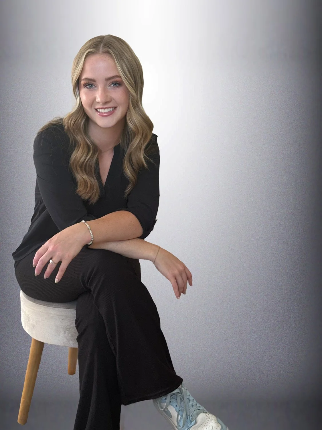 A young woman with long wavy blonde hair sitting on a stool against a gradient grey background, smiling at the camera, wearing a black top, black pants, and white sneakers.