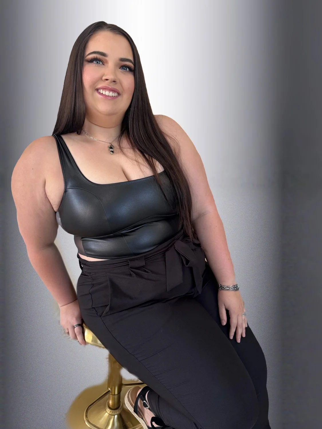 A woman with long brown hair, blue eyes, and light makeup, smiling, wearing a black leather sleeveless top and black pants, sitting on a gold-colored chair against a gray background.