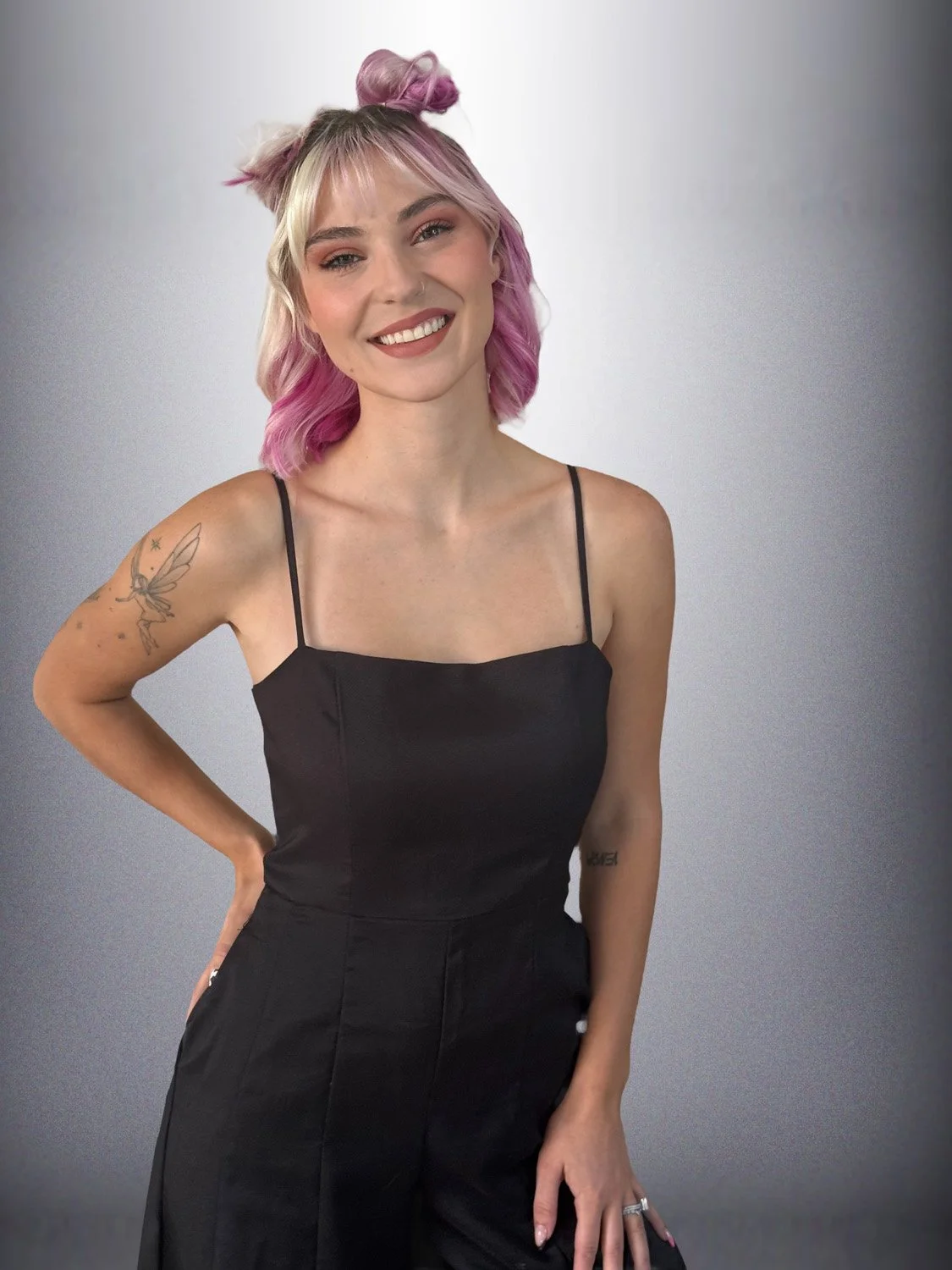 A woman with pink and blonde hair styled in buns and loose waves, wearing a black spaghetti strap top and matching pants, standing against a grey gradient background, smiling.