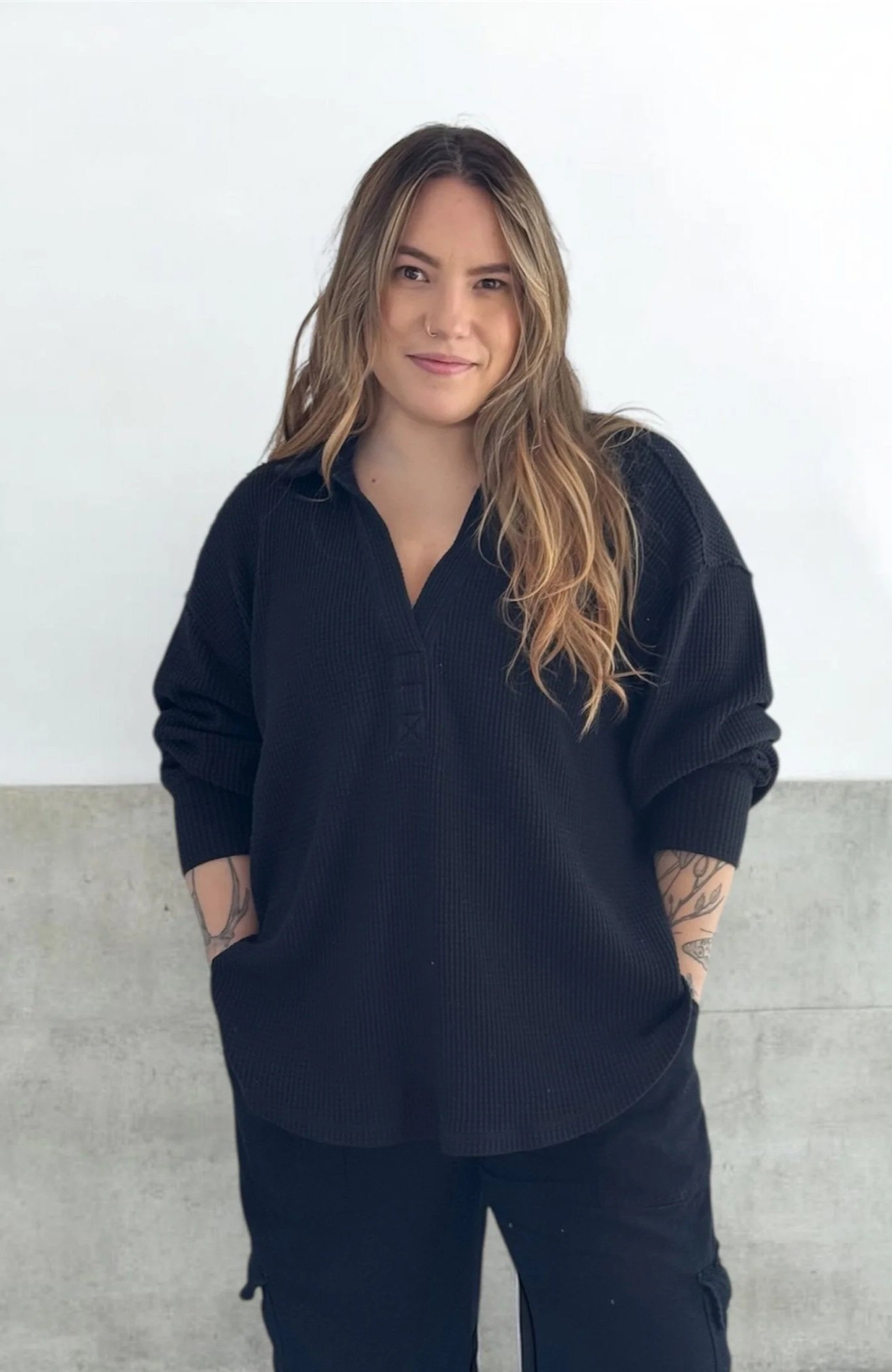 A young woman with long wavy hair and tattoos on her arms, wearing a black oversized sweater, standing against a white wall with a concrete-like lower section, smiling slightly.