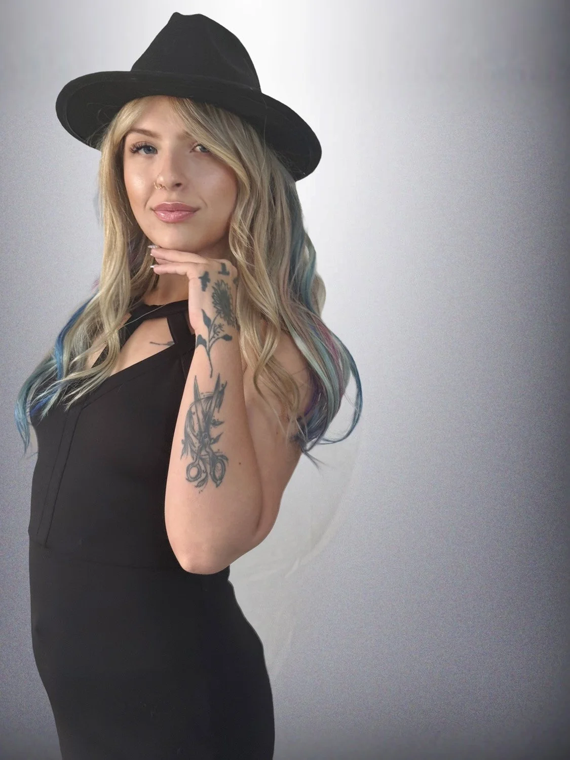 A young woman with long blonde hair with colorful streaks, wearing a black wide-brimmed hat and a black sleeveless dress, poses with her hand under her chin against a simple background.
