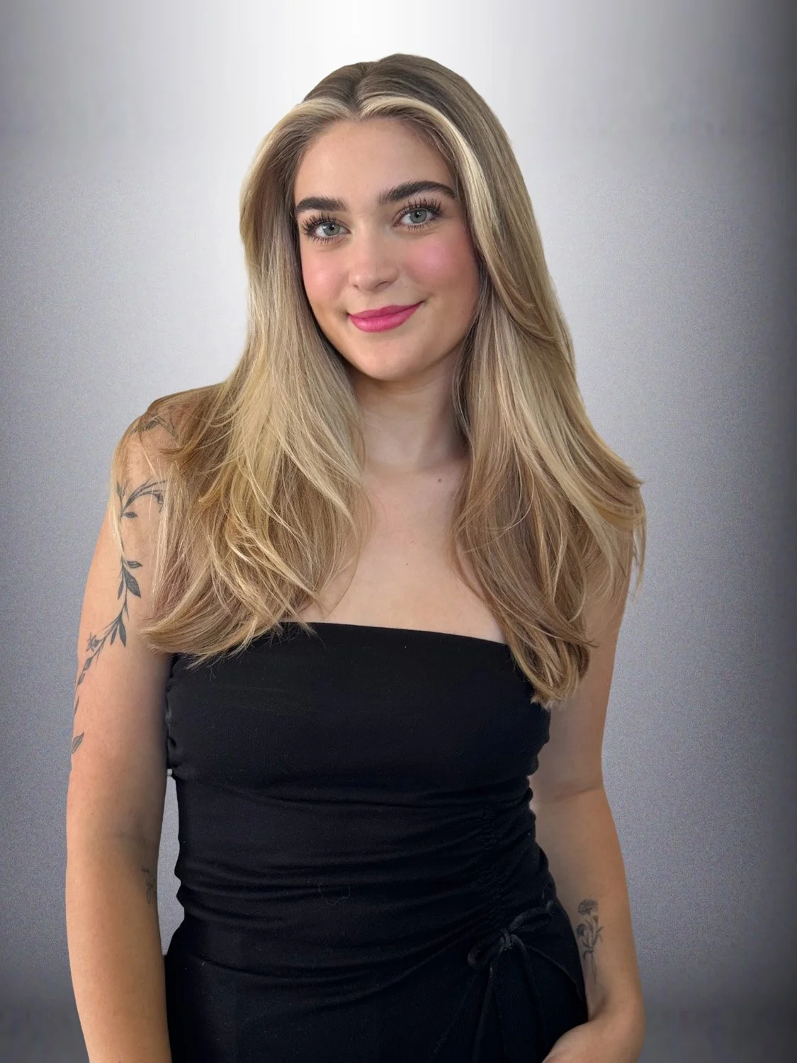 A young woman with long, blonde hair, blue eyes, and makeup, wearing a strapless black dress with tattoos on her arms, smiling at the camera against a plain background.