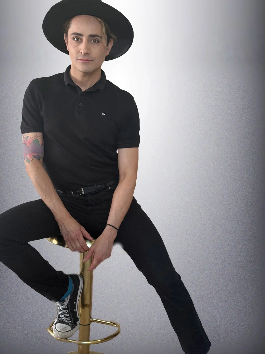 A person with a piercing and a tattoo on their left arm, wearing a black wide-brimmed hat and black clothing, sitting on a gold stool against a plain light gray background.