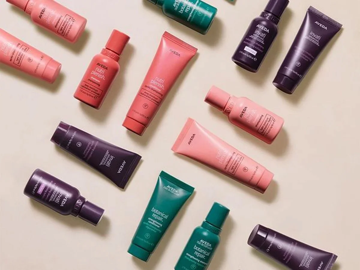 Assorted Aveda hair care products including bottles and tubes in pink, purple, and teal colors displayed on a beige background.