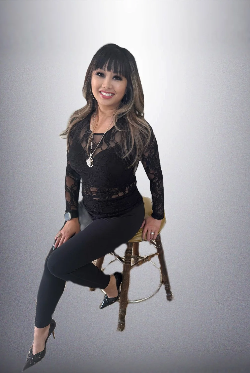 A woman with long wavy hair and bangs, smiling, sitting on a wooden chair with a yellow seat, wearing a black lace top, black pants, black high heels, and jewelry, against a neutral background.