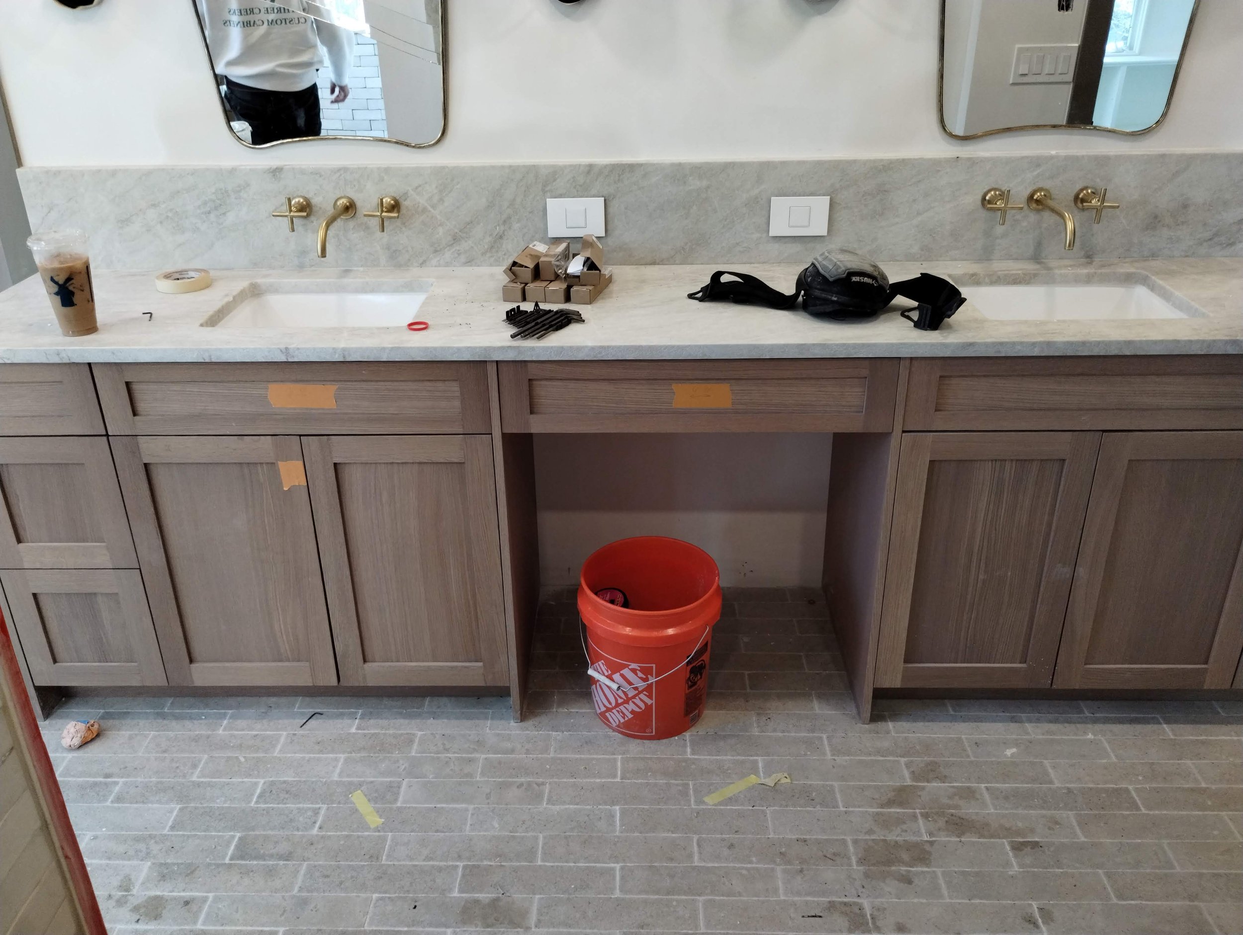 Bathroom under renovation with two sinks, marble countertop, and wooden cabinets. Items include coffee cup, tape, hardware, Home Depot bucket, and work gloves.