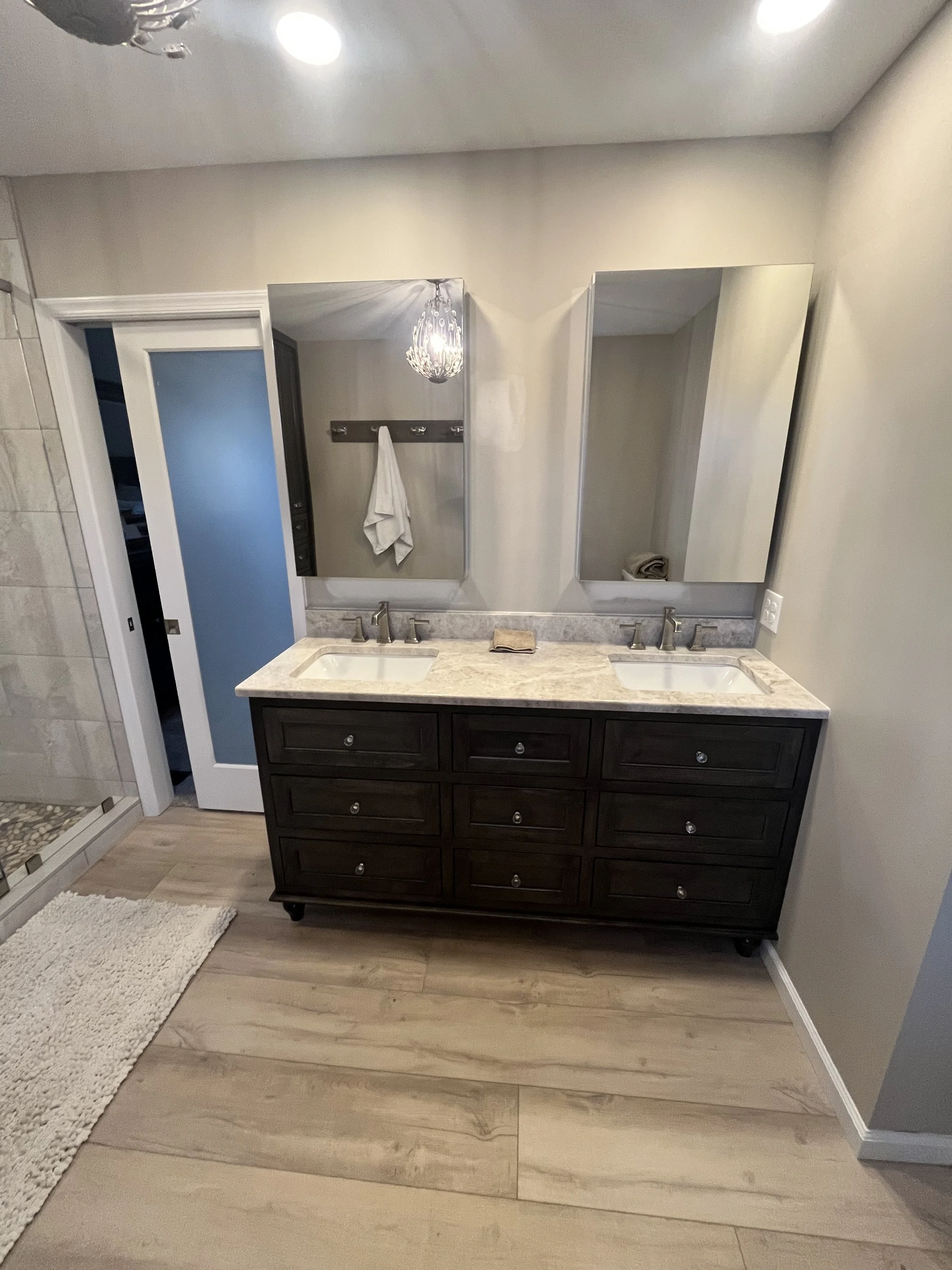 A bathroom vanity with a granite countertop, including two sinks, above which are two mirrors. To the left, a door with a frosted glass panel opens to a shower area with beige tiles. A towel hangs on a hook on the wall, and a small beige rug is on th