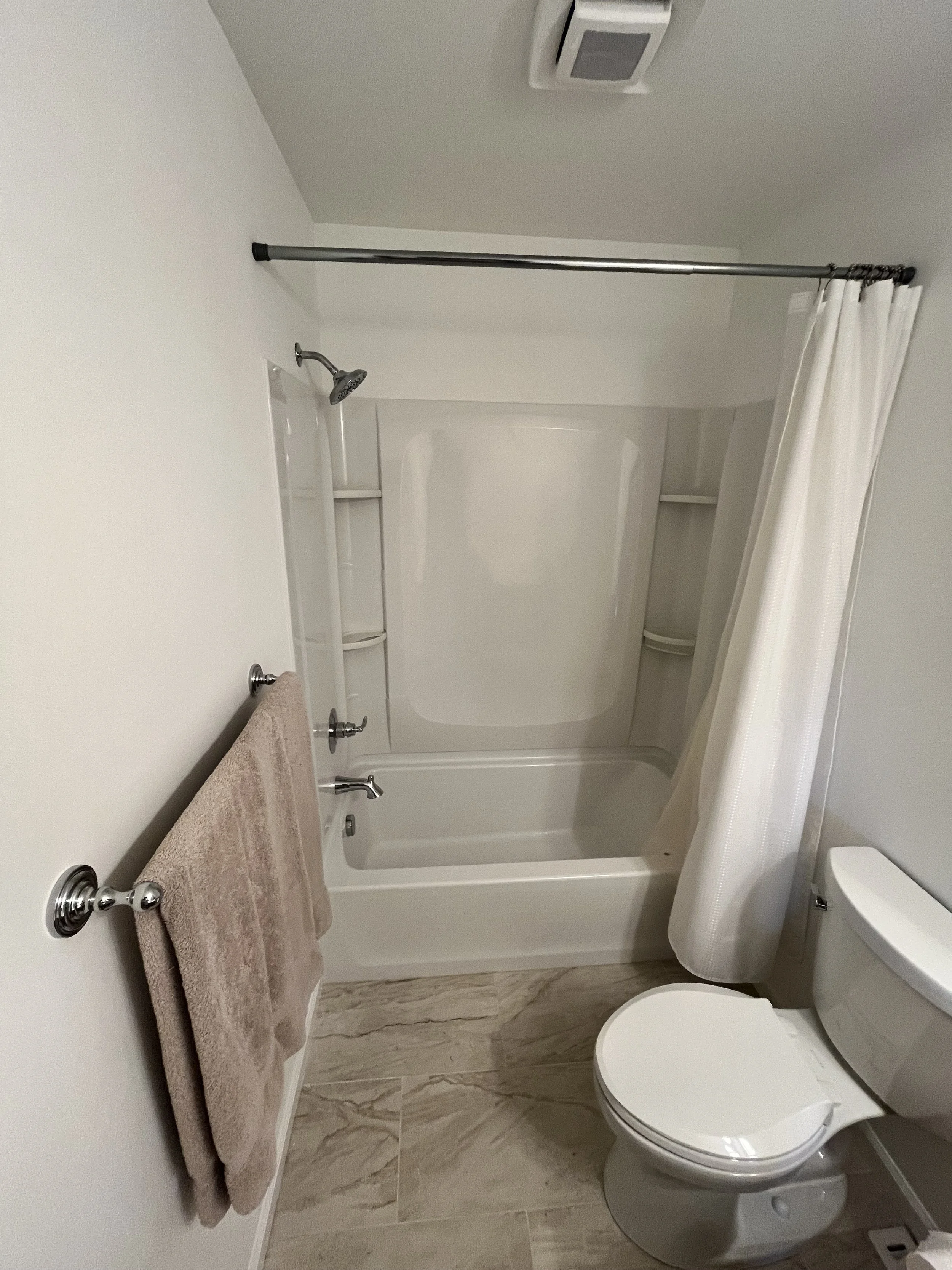 A small bathroom with a shower stall, a toilet, a towel rack with a beige towel, and beige tiled flooring.