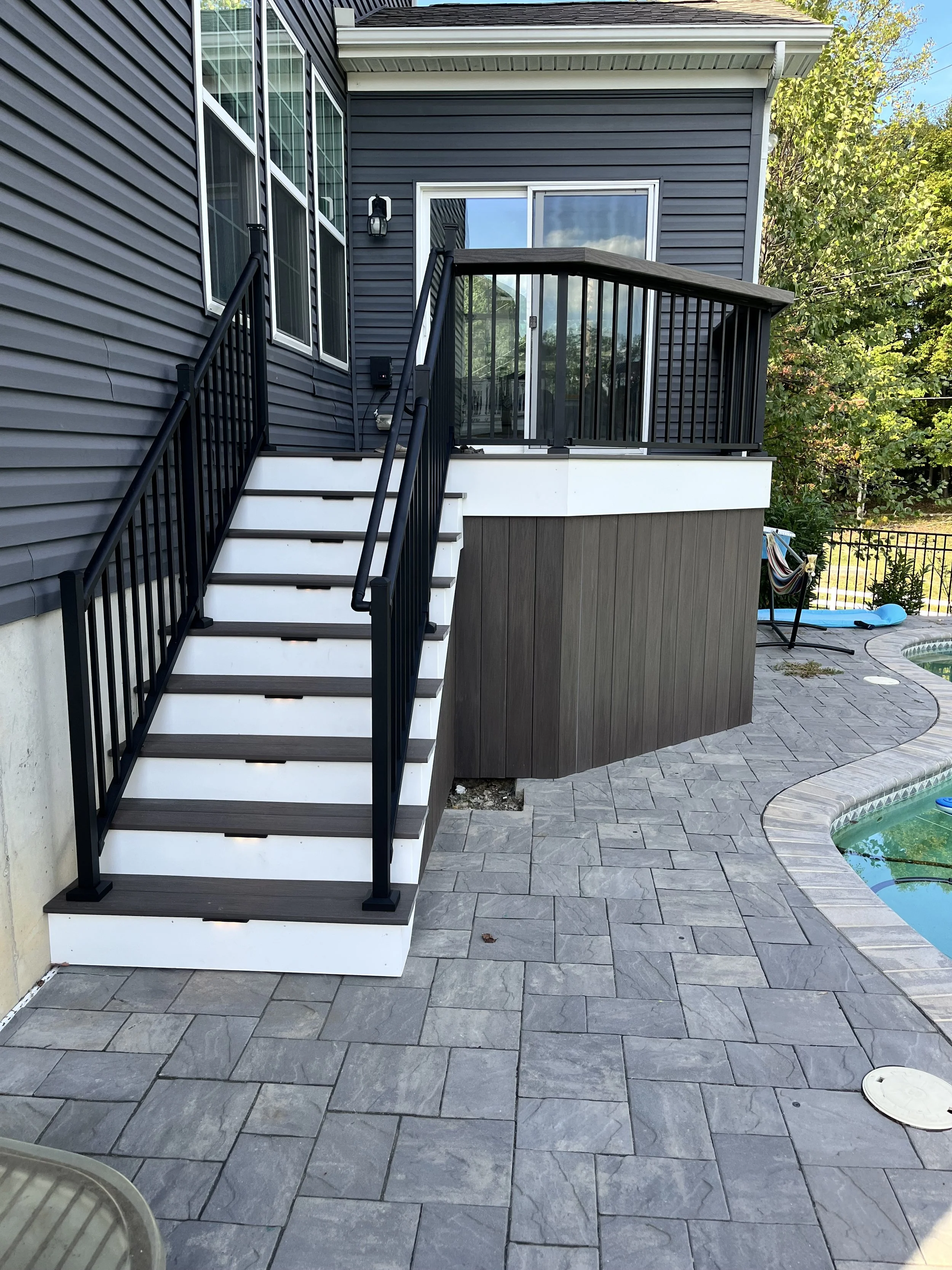 A backyard patio area with grey stone pavers, a small pool, and a set of stairs leading to a house door. The house has blue siding and a fenced yard with trees.