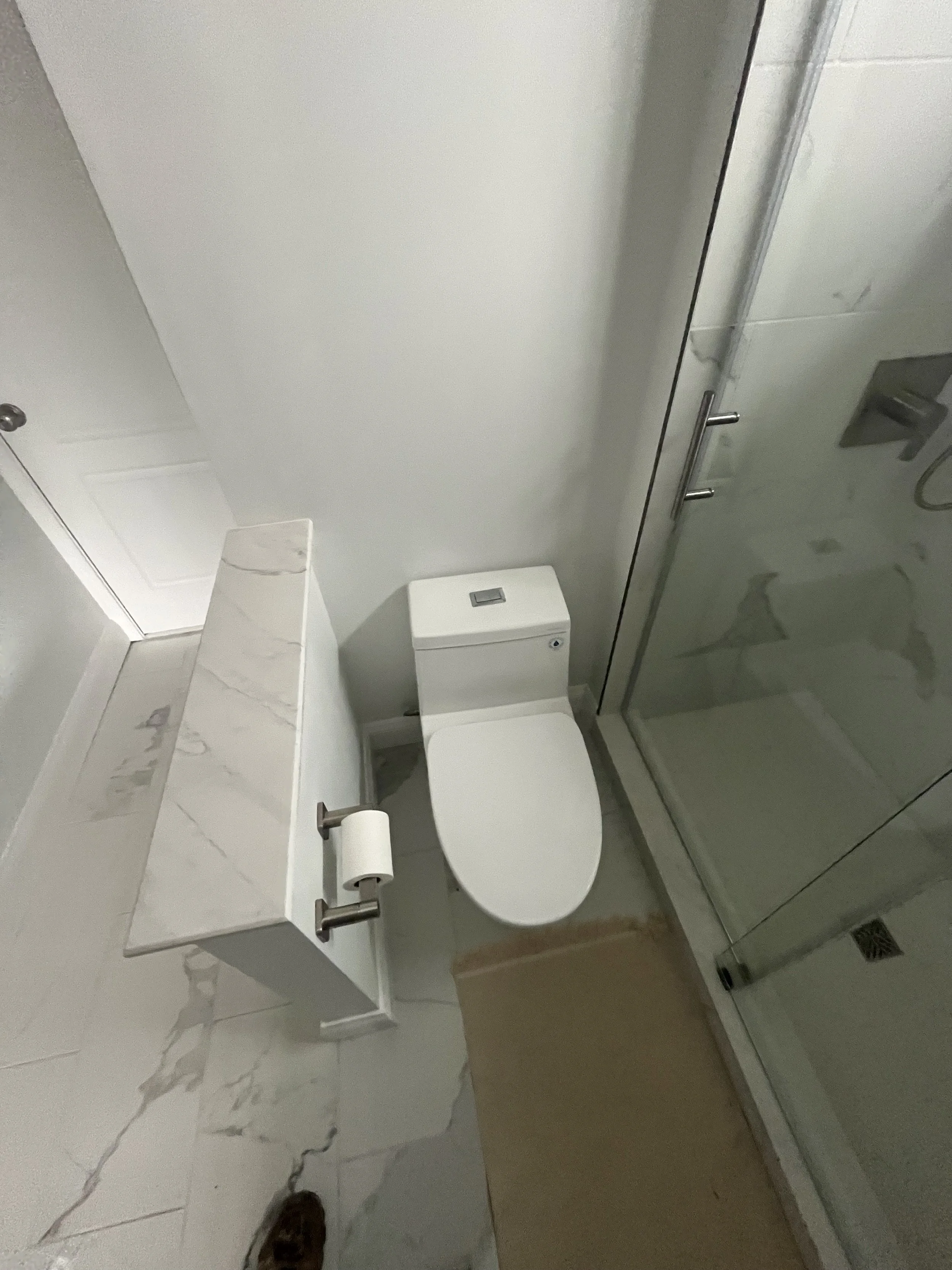 A bathroom with a toilet, a marble countertop with a tissue box, a toilet paper roll, and a shower area with a glass door and marble tiles.