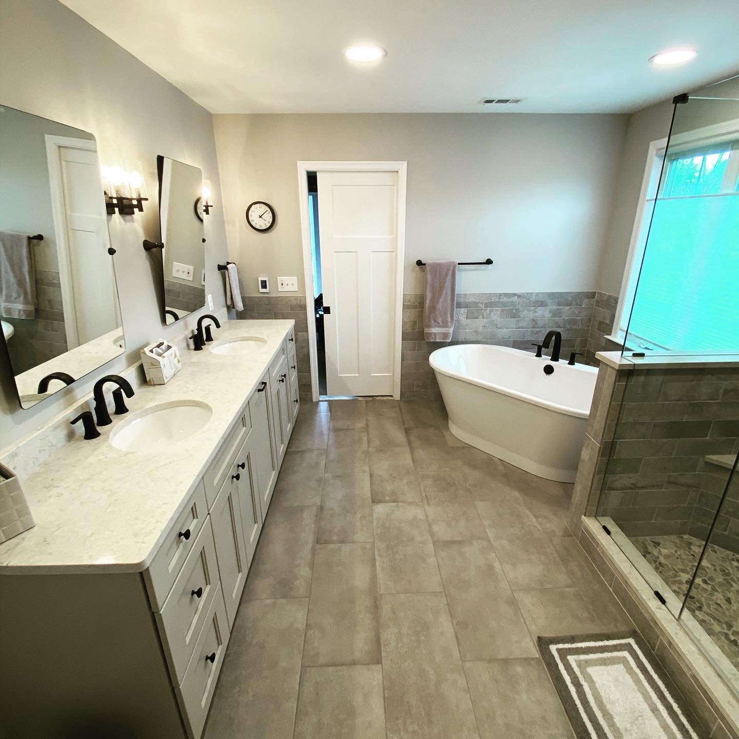 A spacious bathroom with twin sinks on a white vanity, a bathtub, a walk-in shower, and beige tiled floors and walls, illuminated by ceiling lights and natural light from a window.