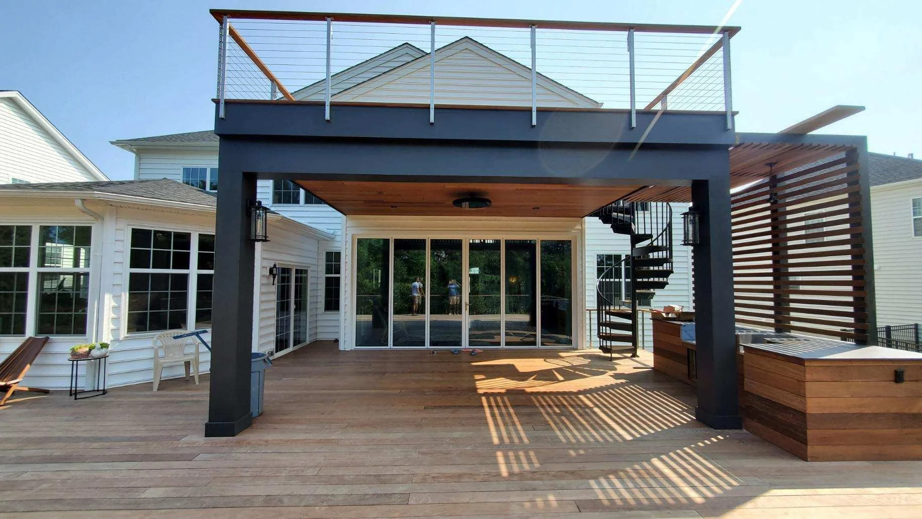 Backyard deck with wooden floor, a covered area with a black roof and railing on top, a spiral staircase on the right, glass sliding doors in the center, and various outdoor furniture pieces.