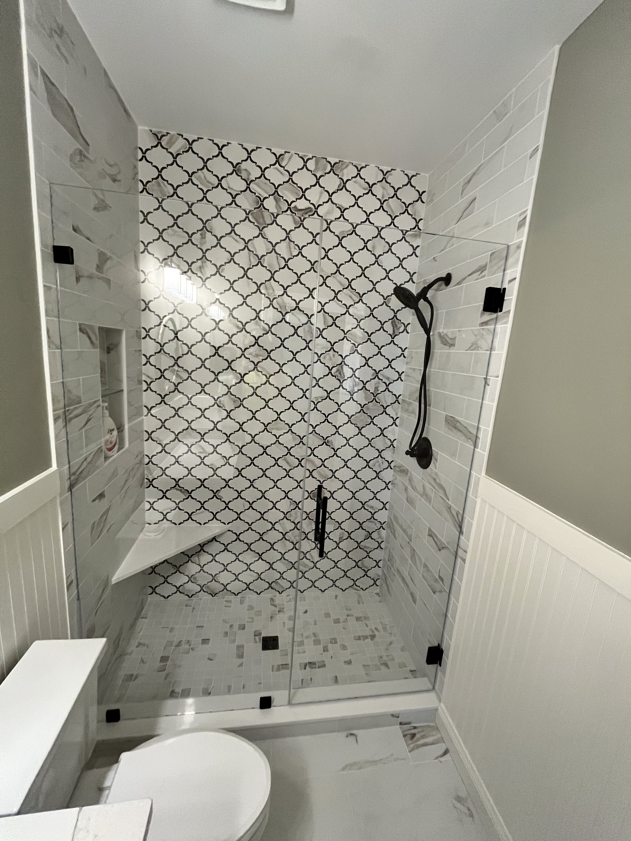 Modern bathroom with a walk-in shower featuring a glass enclosure, black shower fixtures, a patterned tile wall, a built-in shower niche, a toilet, and light-colored marble-like wall tiles.